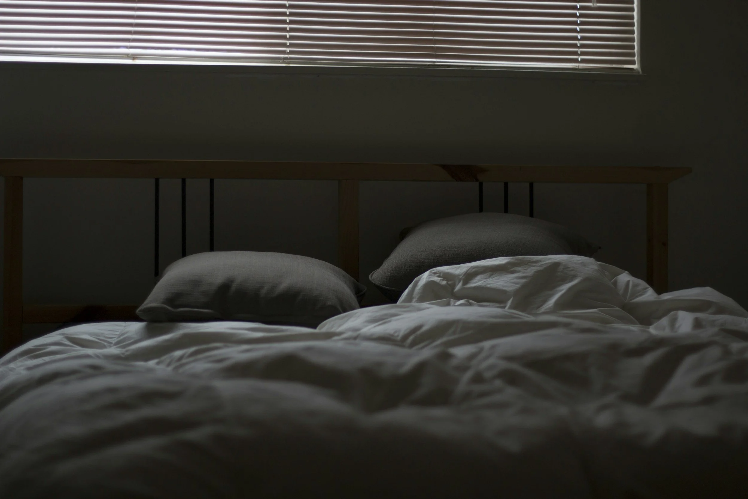 A neatly made bed with white sheets and gray pillows in a bedroom with closed blinds on the window.