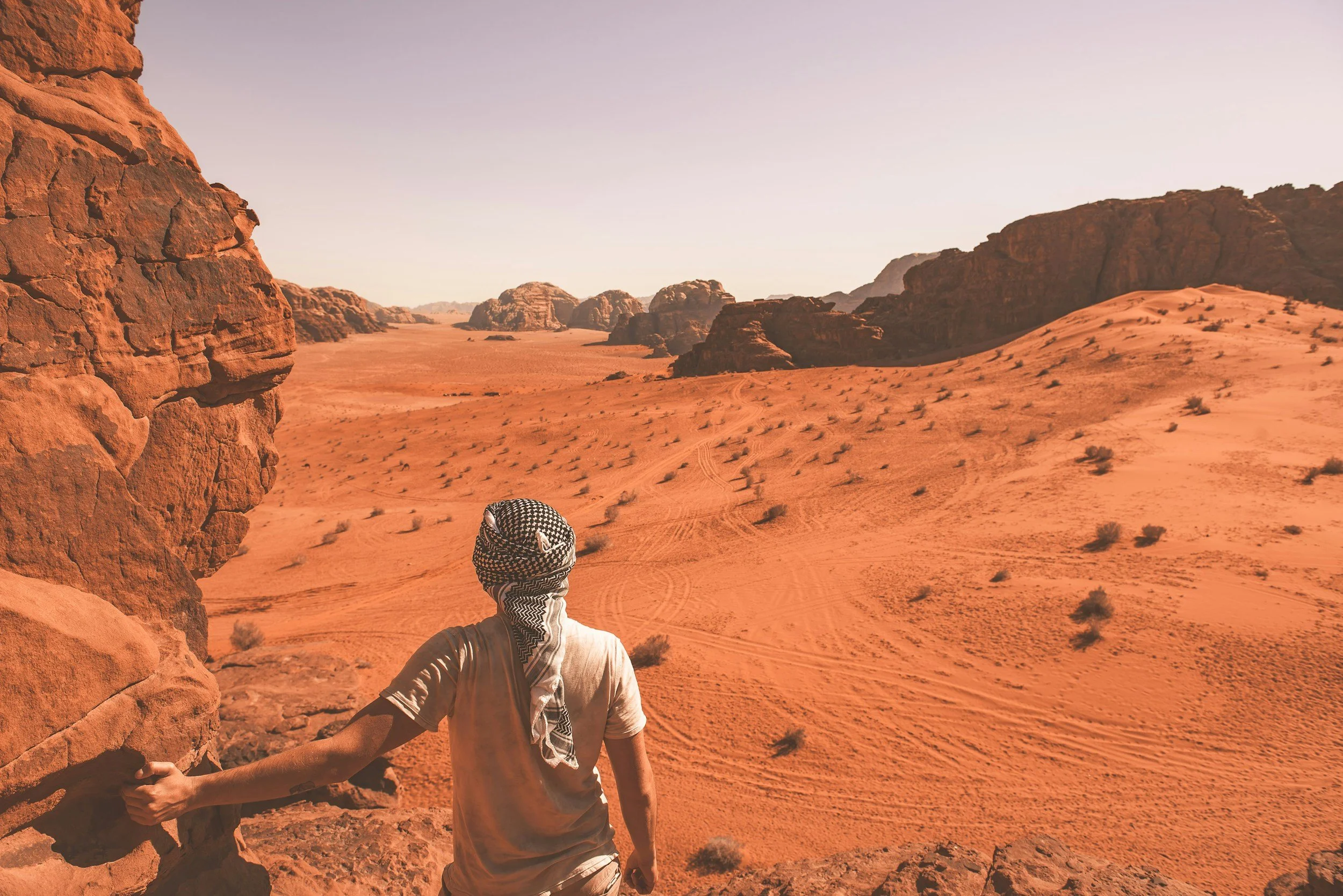 A person in desert terrain wearing a beige shirt and a head covering, holding onto a rock wall, with a vast expanse of red sand, small bushes, and rocky mesas under a clear sky.
