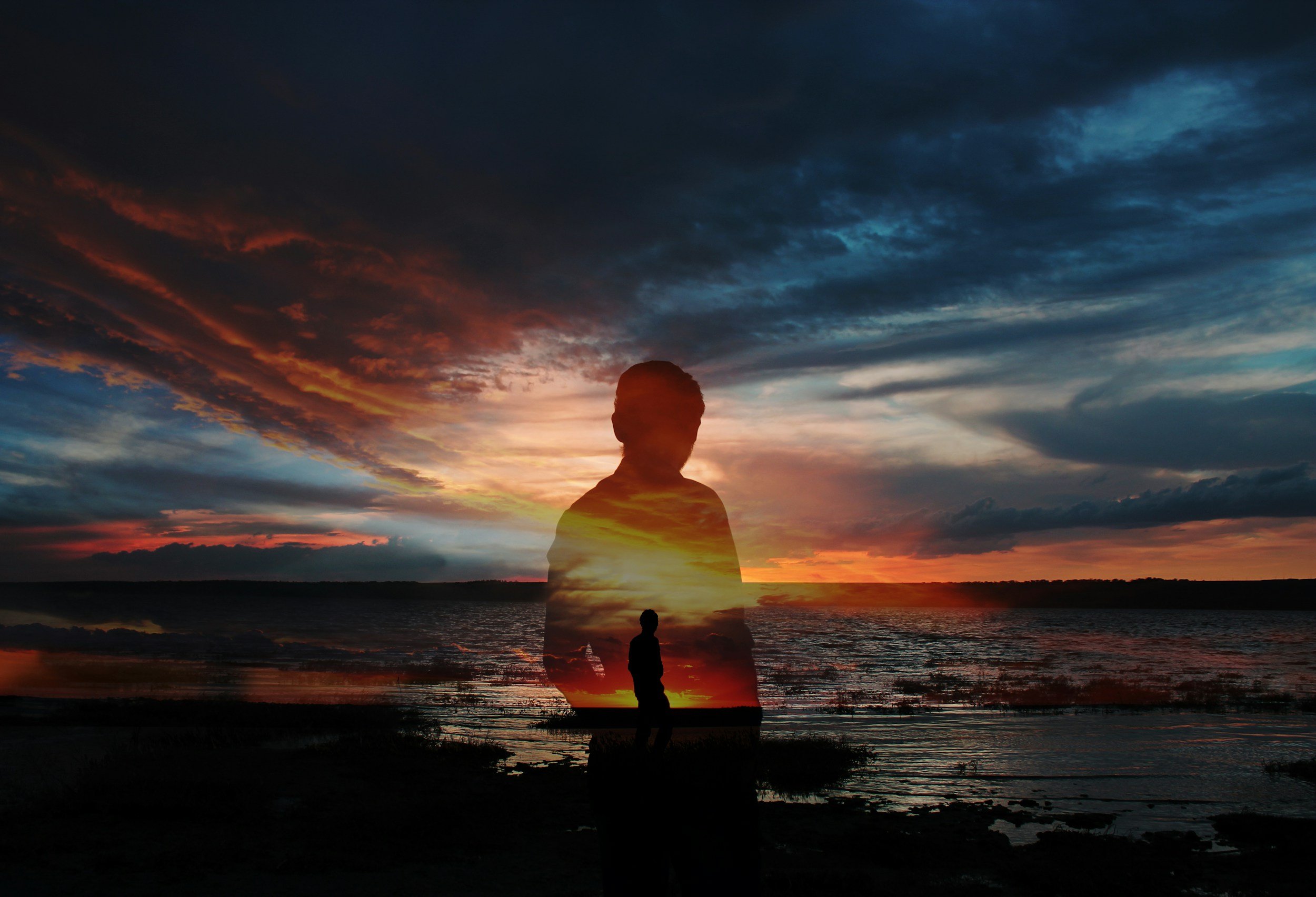 Silhouette of a person standing by the water at sunset with vibrant, colorful clouds overhead.