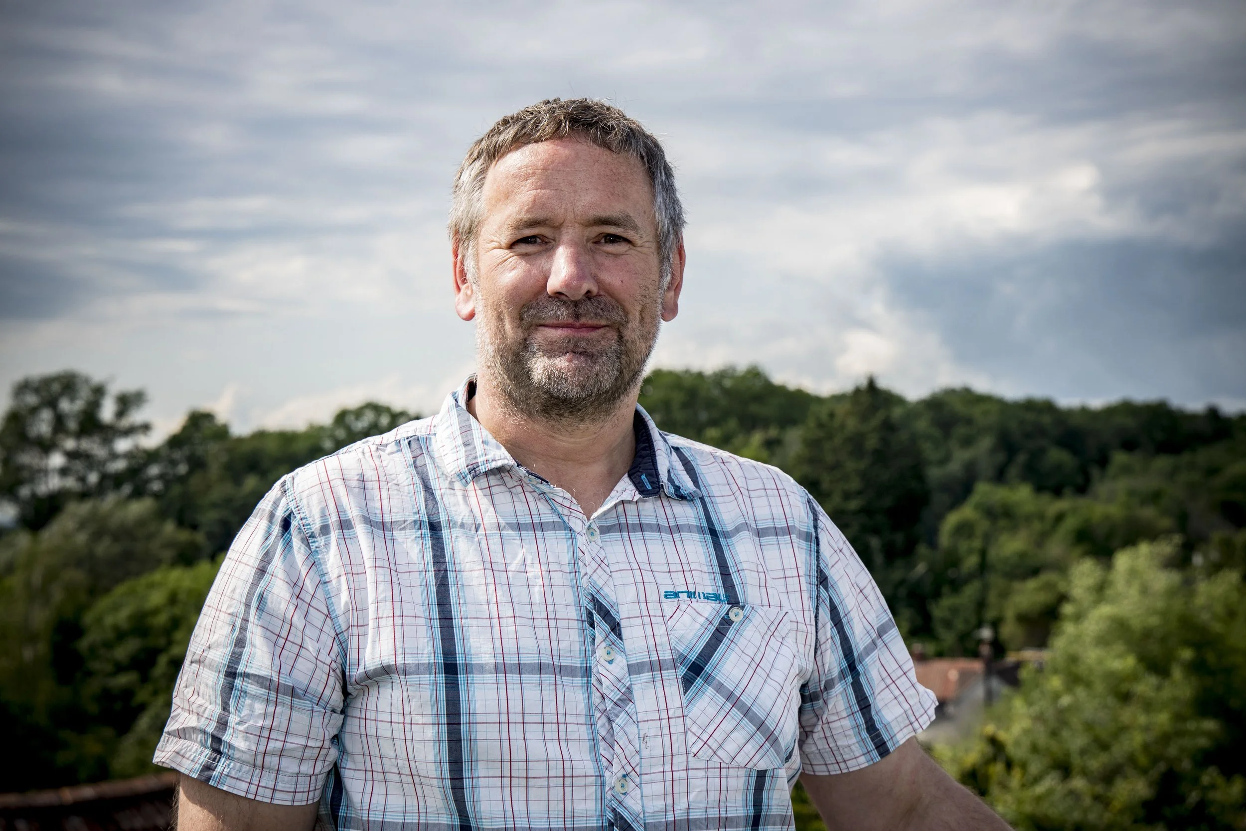 A middle-aged man with gray hair and beard, wearing a light plaid shirt, smiling outdoors with a scenic background of trees and cloudy sky.