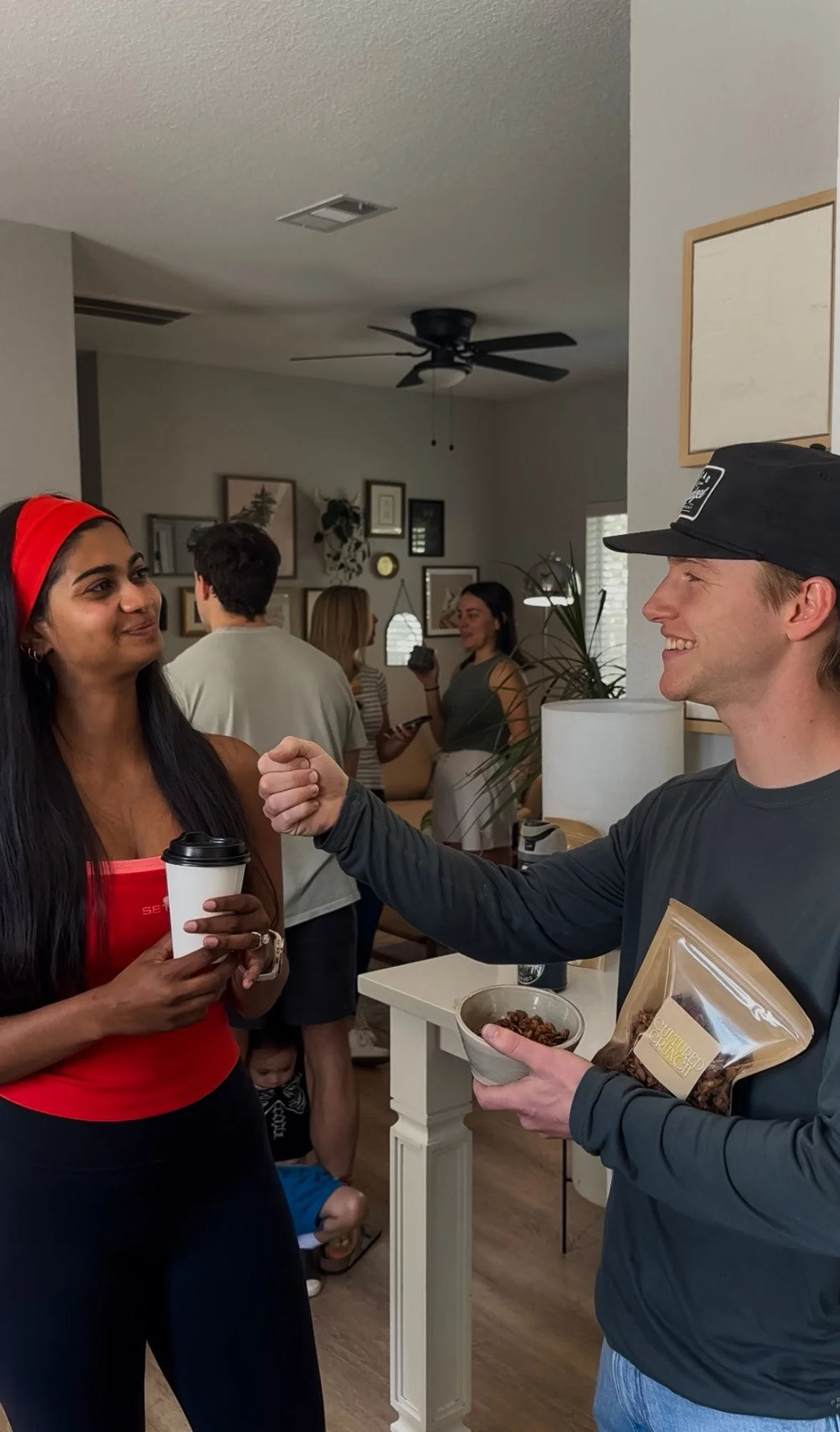 A woman with dark hair and a red headband holds a coffee cup while smiling at a man wearing a black cap. They are indoors, engaged in conversation, with a group of people in the background.