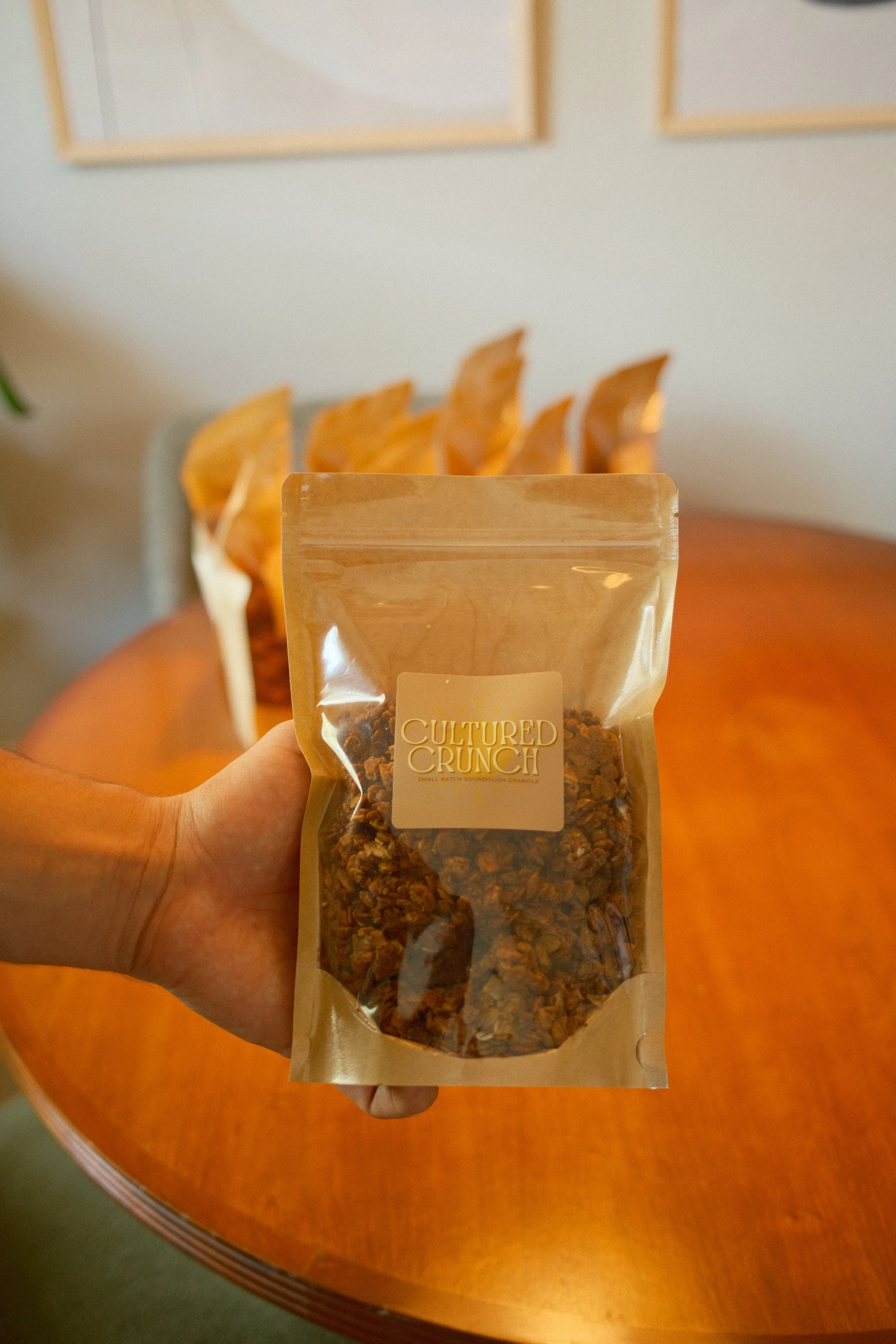 A hand holding a transparent resealable pouch labeled 'Cultured Crunch' with granola inside, in front of a wooden table and blurred background.