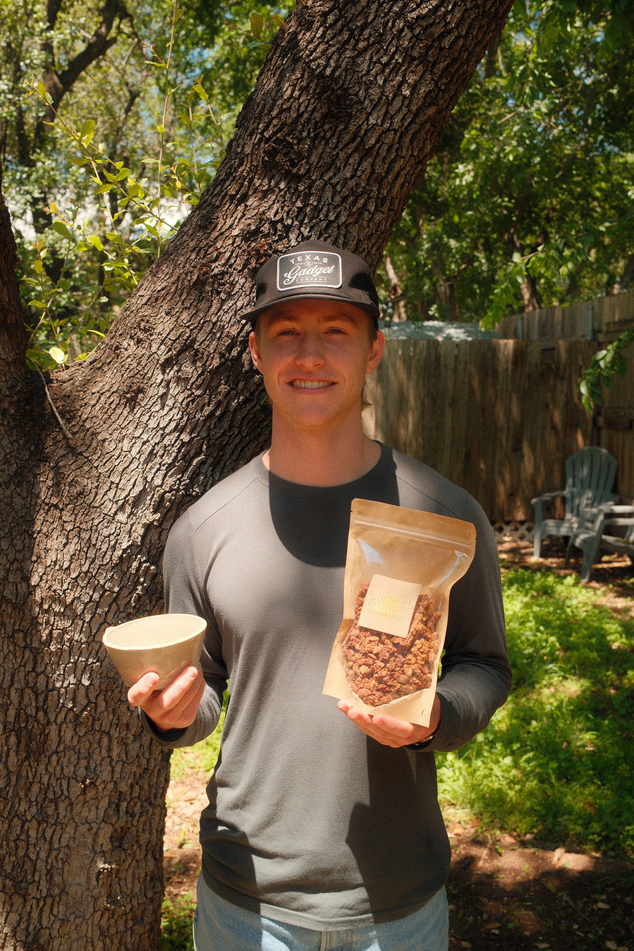 A young man with a black cap, gray and black shirt, smiling and holding a bag of chocolate chip cookies and a beige bowl outdoors near a large tree in a backyard.