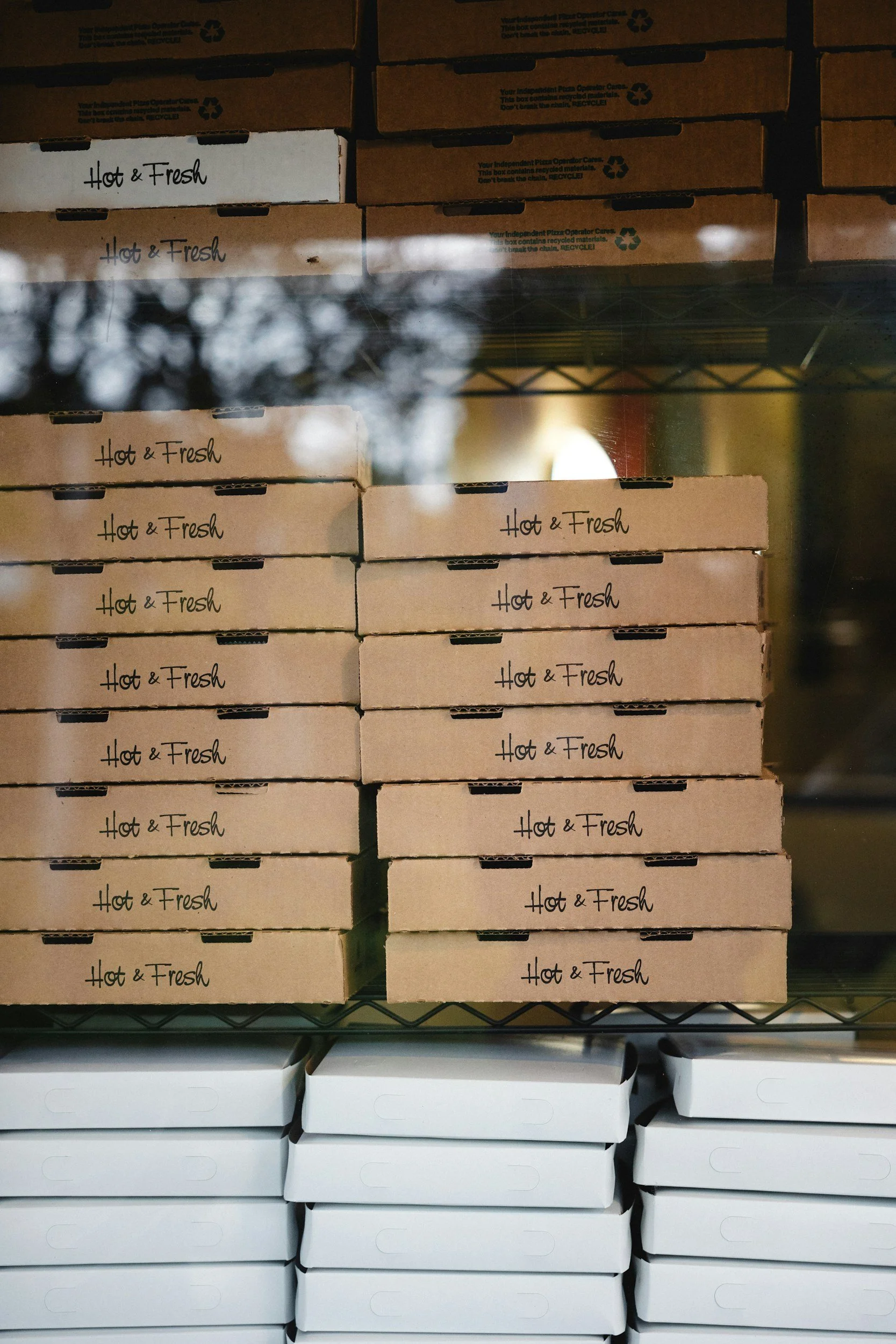 Stacked pizza boxes labeled 'Hot & Fresh' on a shelf, with a reflection of trees visible on the glass door.