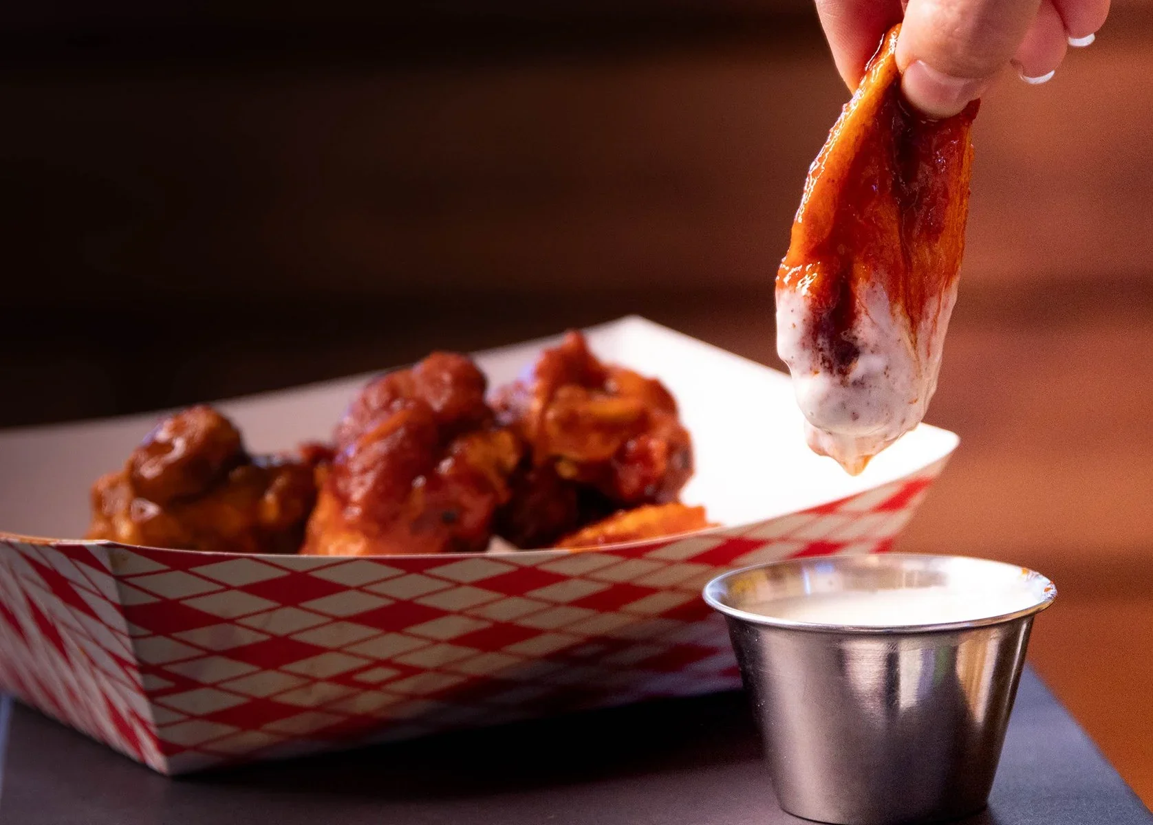 Close-up of buffalo chicken wings in a paper tray with a hand dipping a wing into ranch dressing, with a metal container of ranch on the side.