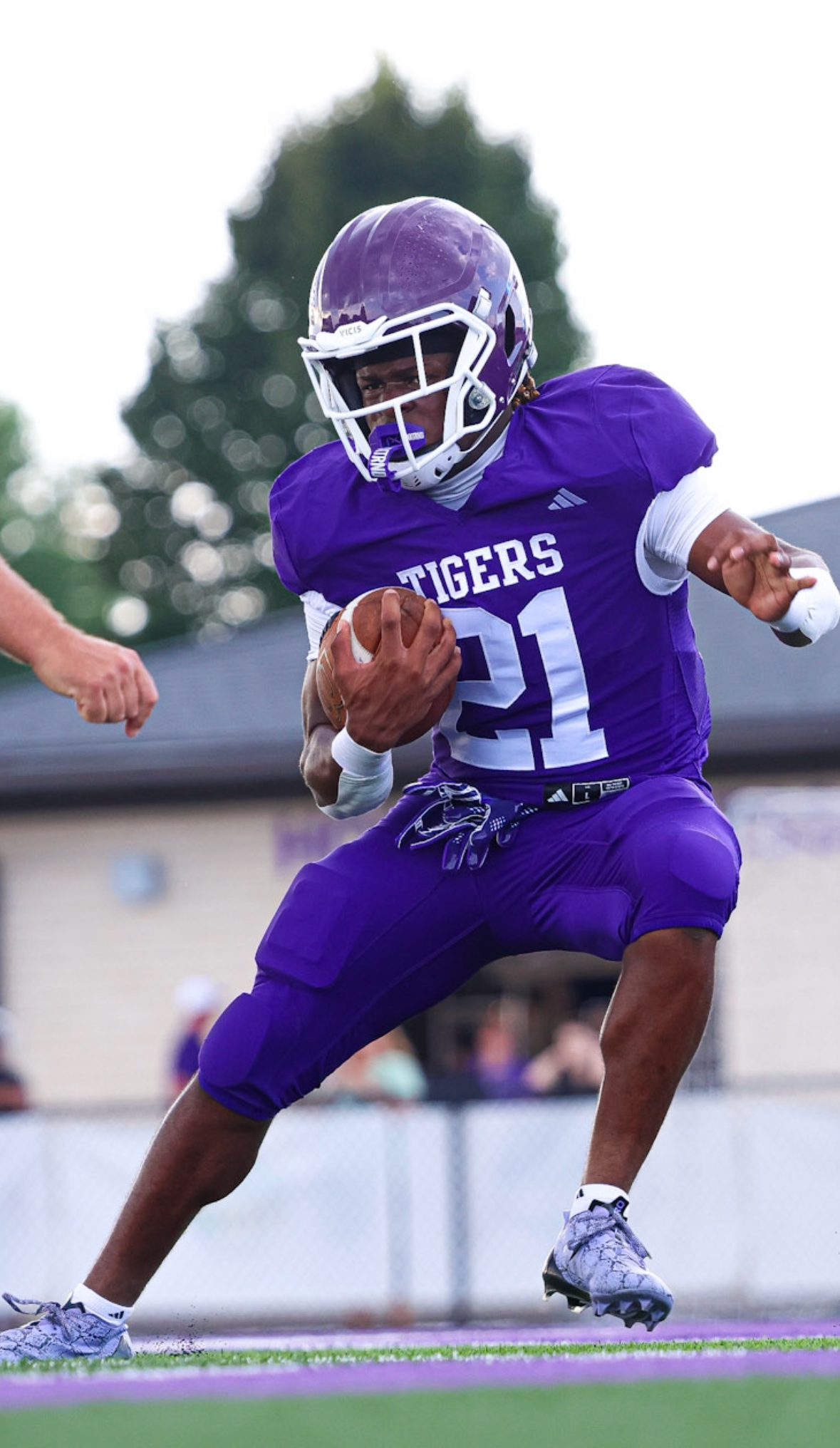 A football player in a purple uniform and helmet, holding a football, appears to be running during a game.