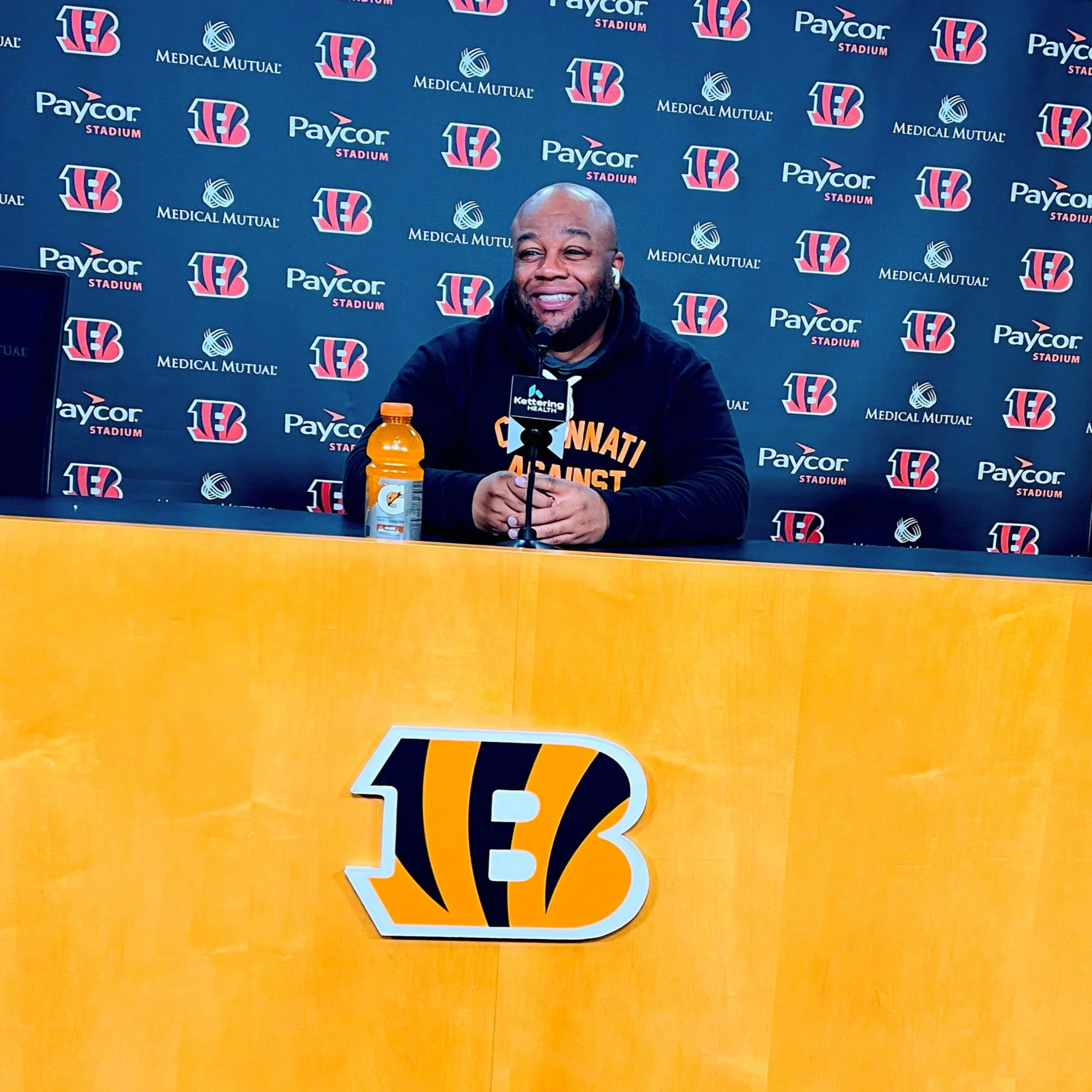 A man smiling during a press conference at Paycor Stadium, with a Cincinnati Bengals themed background and a large Bengals logo on the front of a yellow table. There is an orange Gatorade bottle on the table and a microphone in front of him.