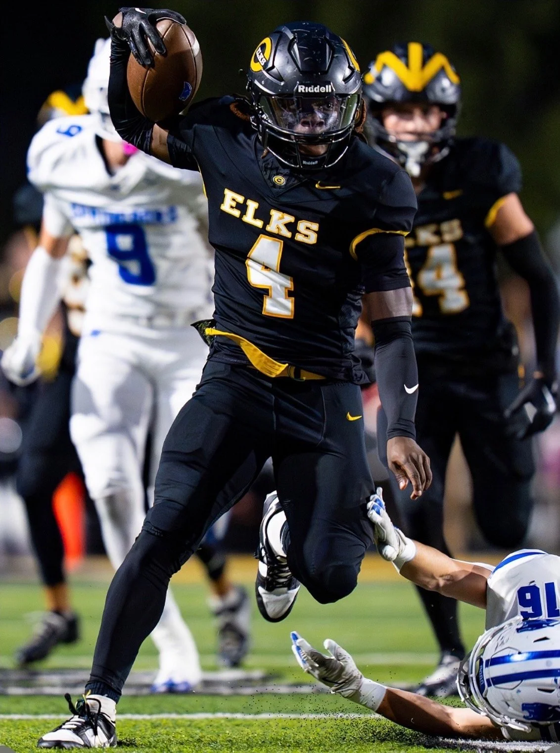 A football player wearing a black uniform with yellow accents, black helmet, and gloves jumping with the ball in hand, surrounded by opponents on the field during a game.