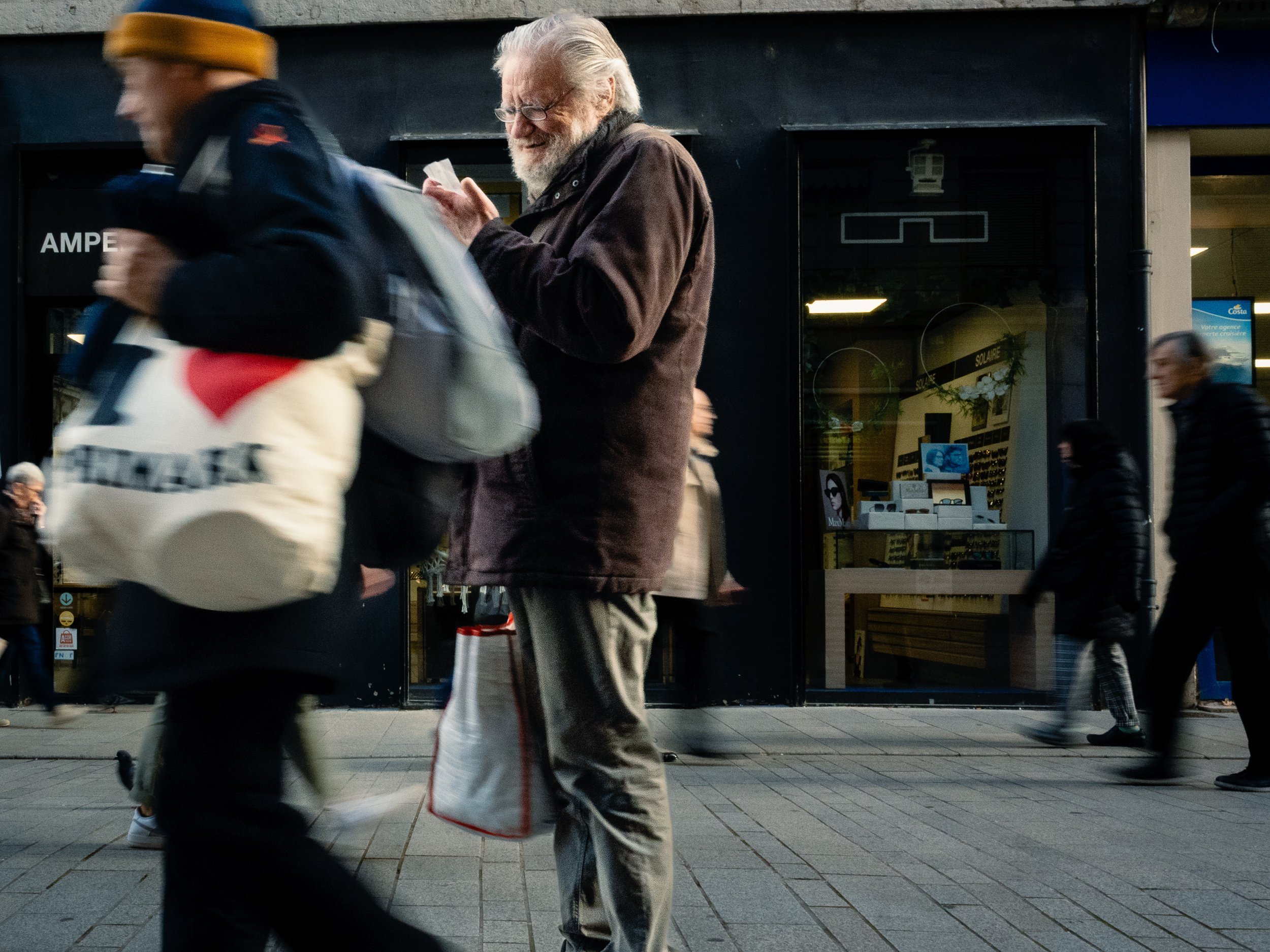 People walking past a storefront with large windows. An elderly man with white hair and a beard is visible.  He's reading a slip of paper and wearing glasses. A blurred figure in the foreground is and another person is walking by in the background. 