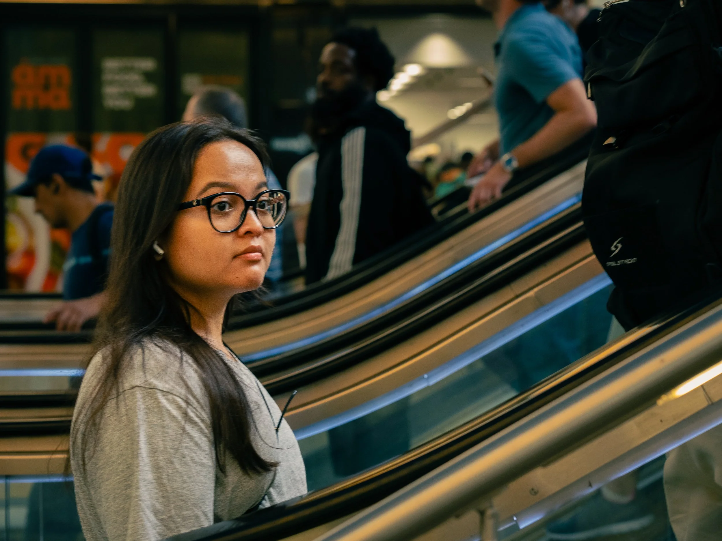 A woman with long dark hair and glasses standing on an escalator in an indoor public space with other people around.