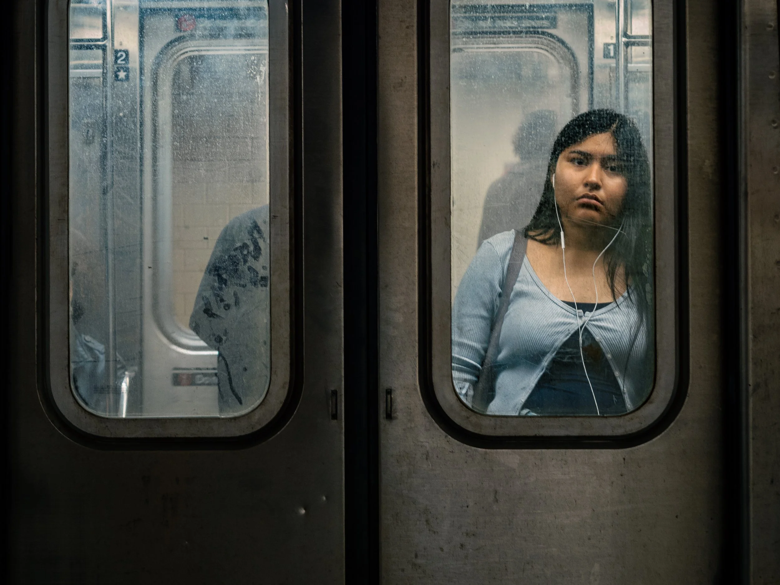 A young woman with dark hair, wearing earphones and a light blue top, looking out a foggy subway train window.