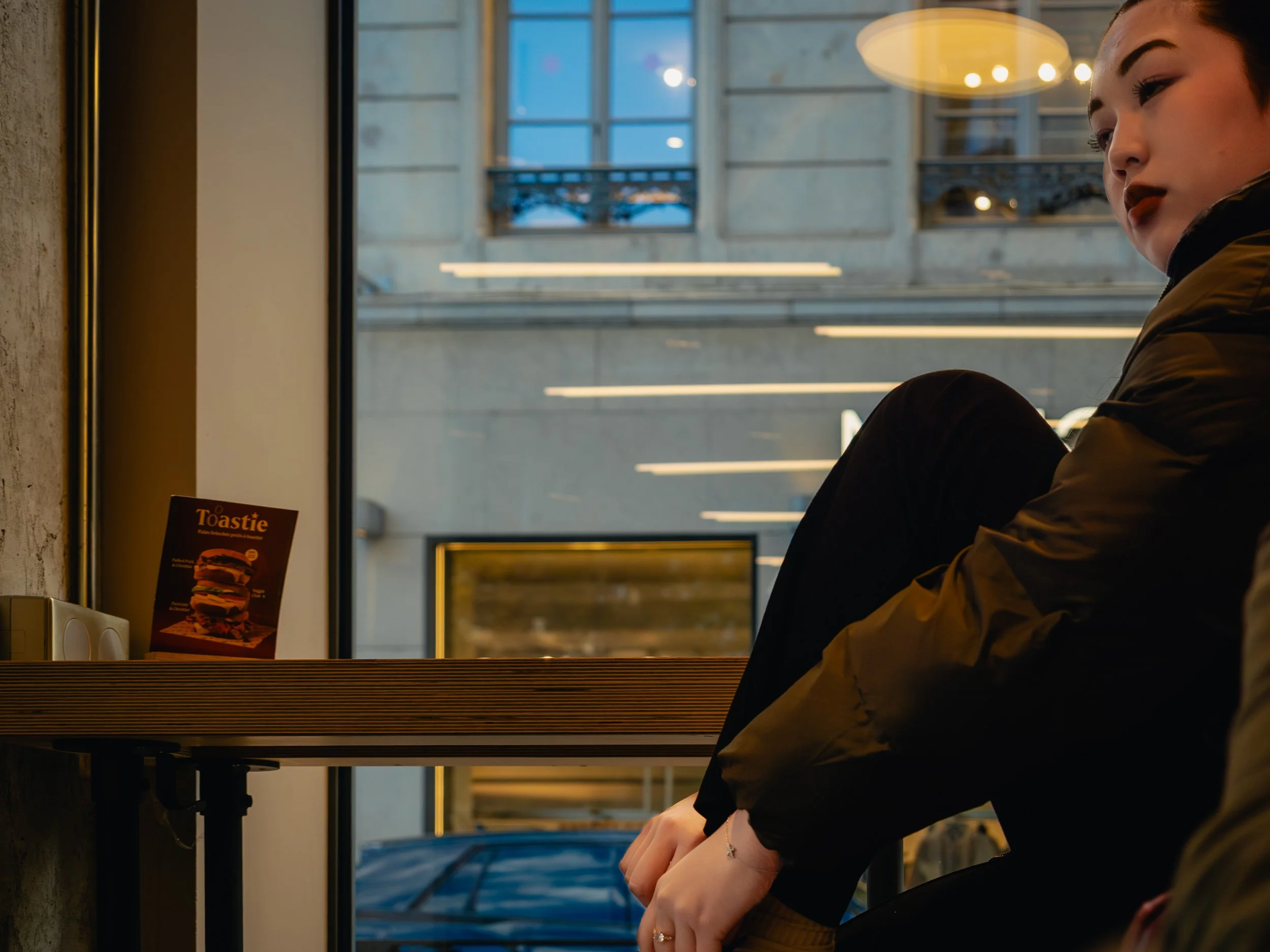 A person with dark hair and makeup sitting by a window in a modern building, wearing a jacket, with a poster of a burger on the table next to them.