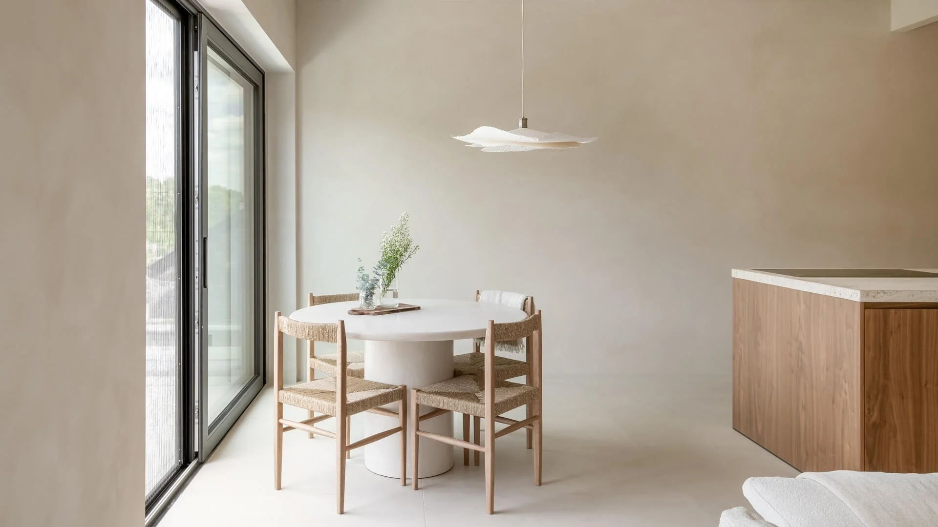 Minimalist dining area with a white round table and four wooden chairs with woven seats, situated next to large sliding glass doors leading outside. A modern, white pendant light hangs above the table, and a small plant in a glass vase is on the table. Part of a kitchen countertop with a wood finish is visible on the right.