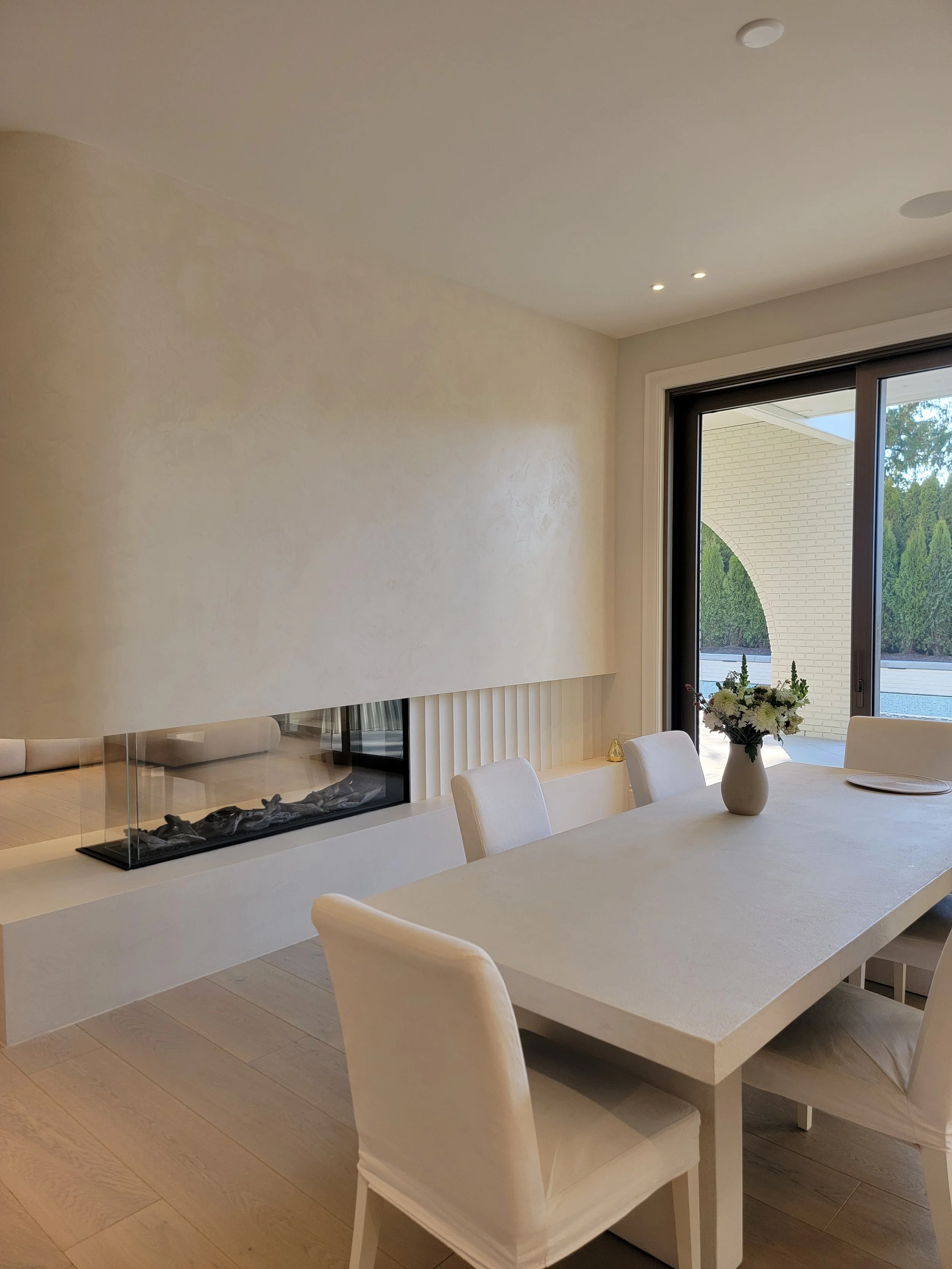 Modern dining room with white table and chairs, fireplace with glass enclosure, large window with view of greenery outside, potted flowers on table.