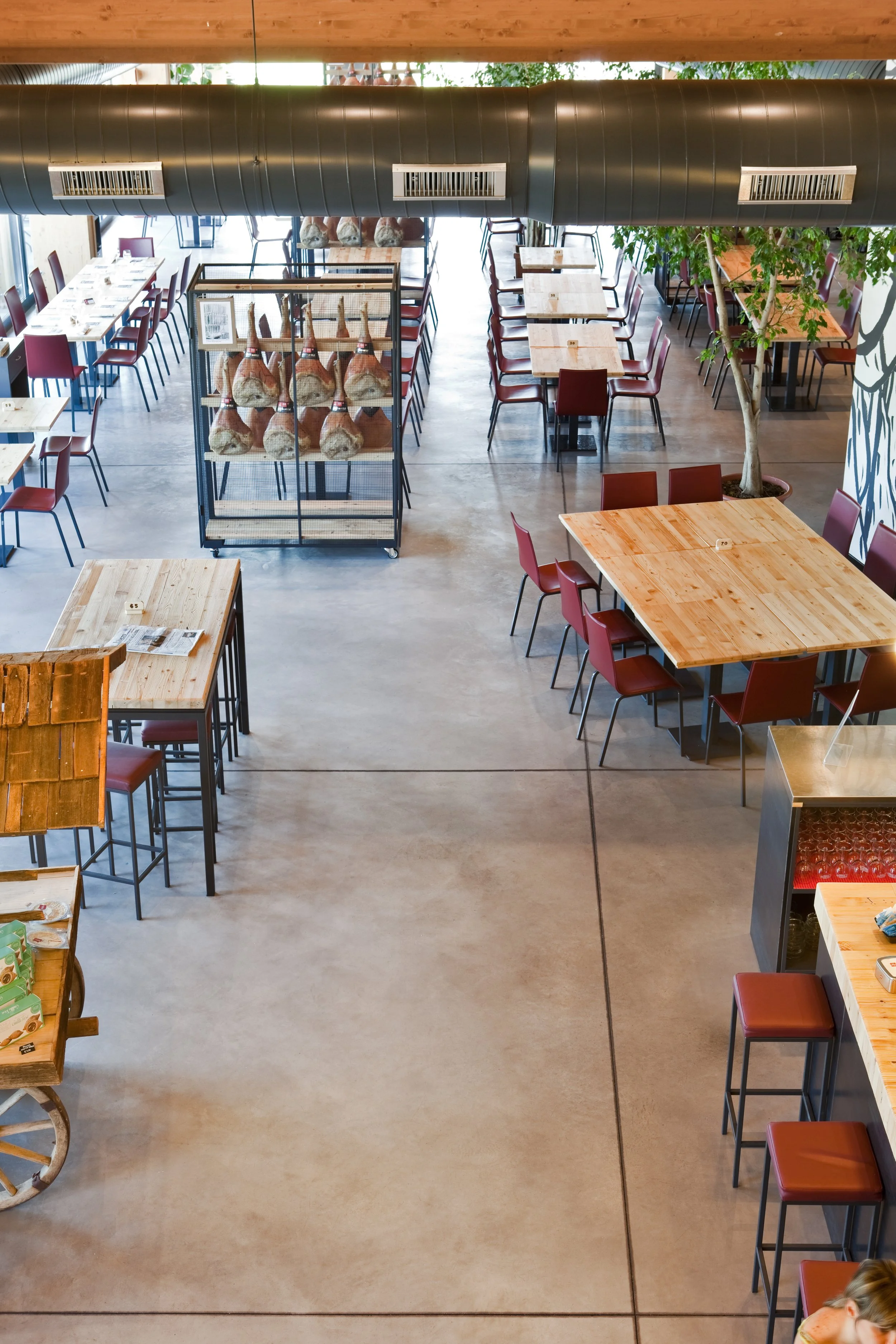Interior of a restaurant or cafe with wooden tables and red chairs. There is a large dark grey air duct running across the ceiling. A metal cage displays hanging cured meats. A tall potted tree is positioned near some tables on the right side.