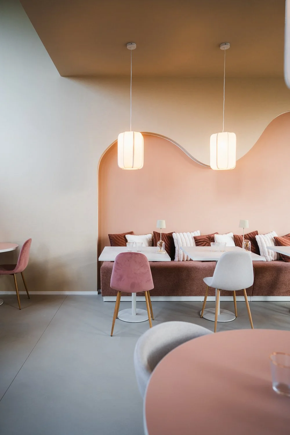 Interior of a modern cafe with pastel pink, white, and brown decor, featuring hanging pendant lights, a pink cushioned banquette with pillows, and small tables with pink and white chairs.