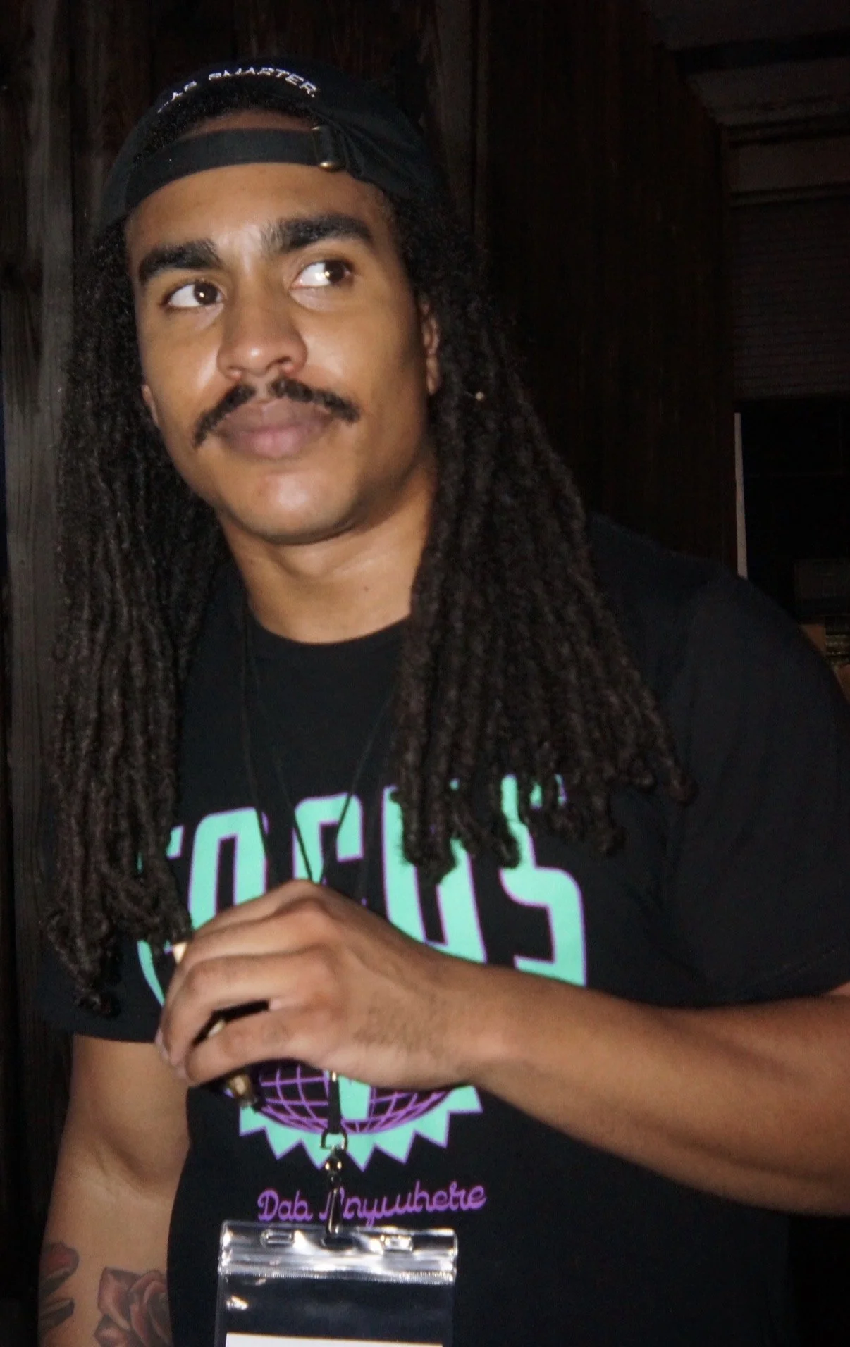 Person with long, dark dreadlocks, wearing a black cap backwards and a black T-shirt with a colorful graphic and text. The person has a mustache and goatee, and is holding a lanyard with a badge. The background is dark with wood paneling.