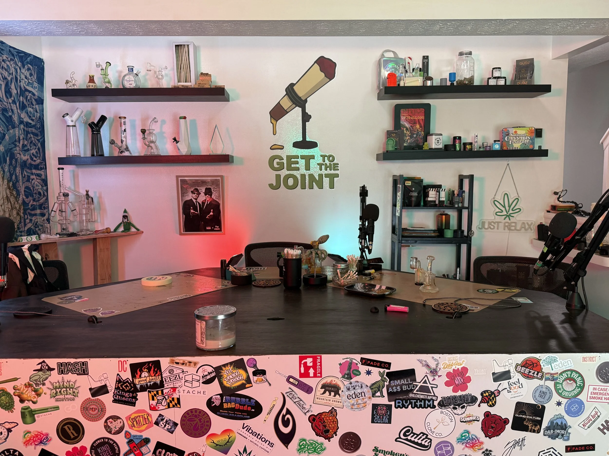 A room with shelves holding glass paraphernalia, books, and cannabis products, a wall sticker of a lit joint with the phrase "Get to the joint," and a table with colorful stickers.