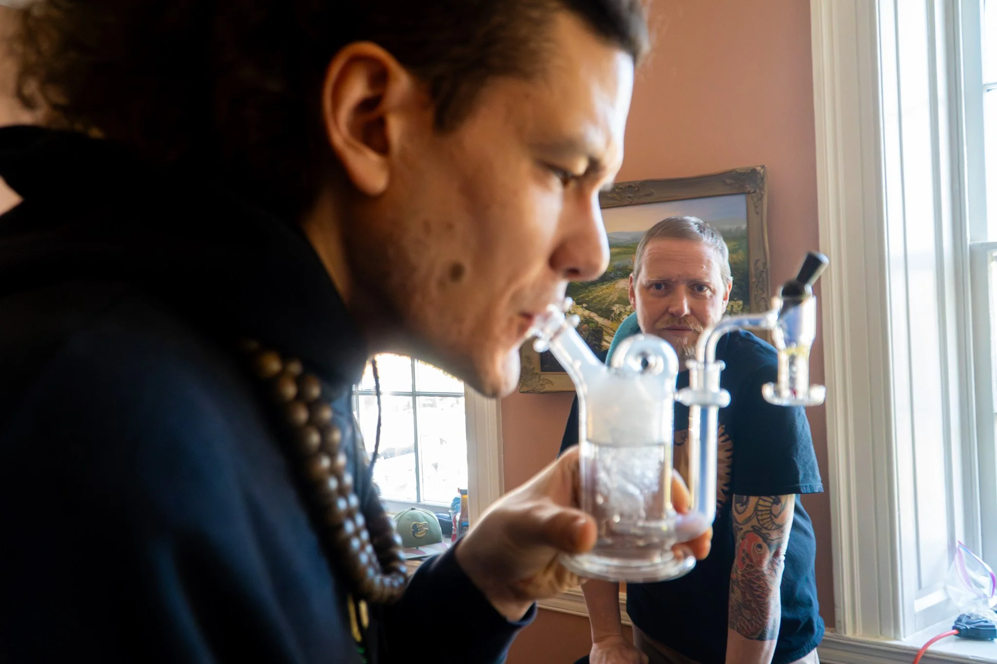 Two men are sitting at a table in a room filled with natural light. The man in the foreground is holding a glass of drink and appears to be looking at something on the table, while the man in the background is observing him. The room has a window with white trim and a landscape painting on the wall.