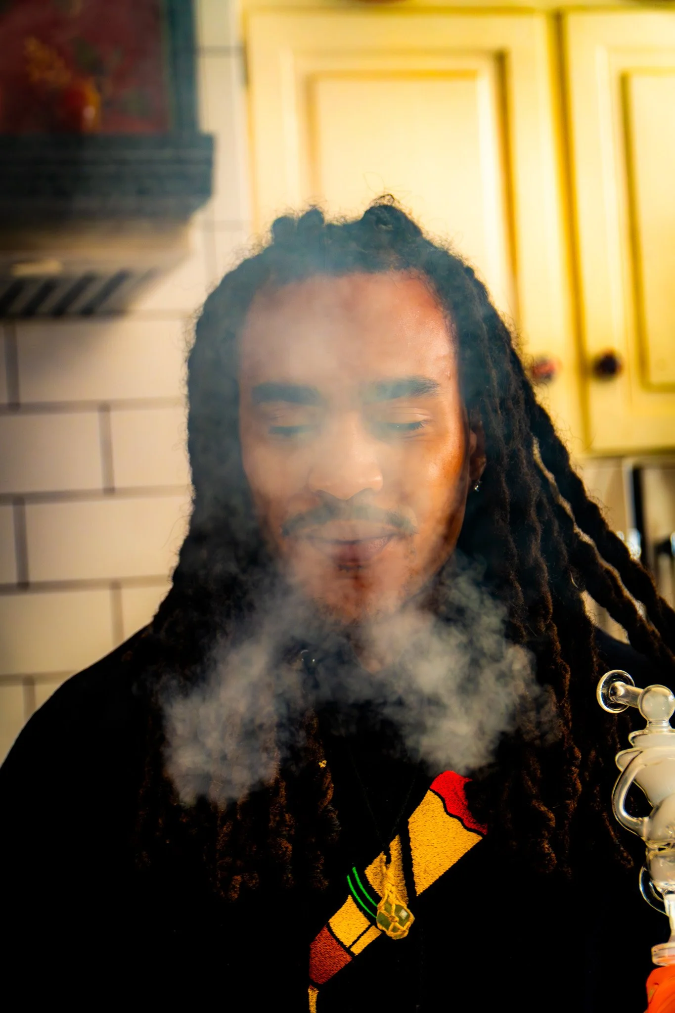 A person with dreadlocks and a dark hoodie, standing in front of a kitchen cabinet, exhaling smoke.