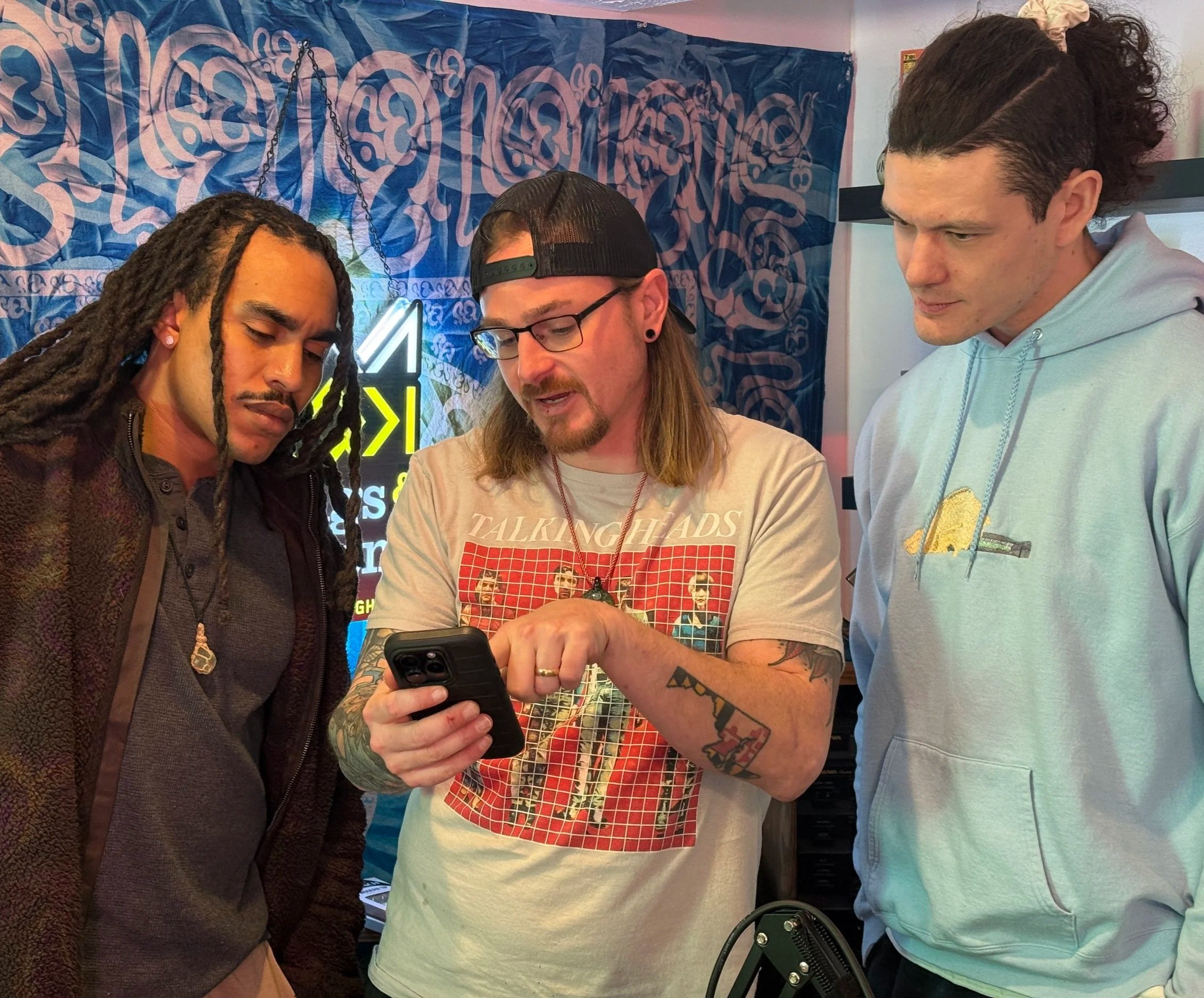 Three men gathered together looking at a smartphone. The man in the center has tattoos, glasses, and a backwards cap. The man on the left has dreadlocks and jewelry. The man on the right has dark, curly hair and is wearing a light blue hoodie. They are in a space with a blue and pink graffiti-style backdrop.