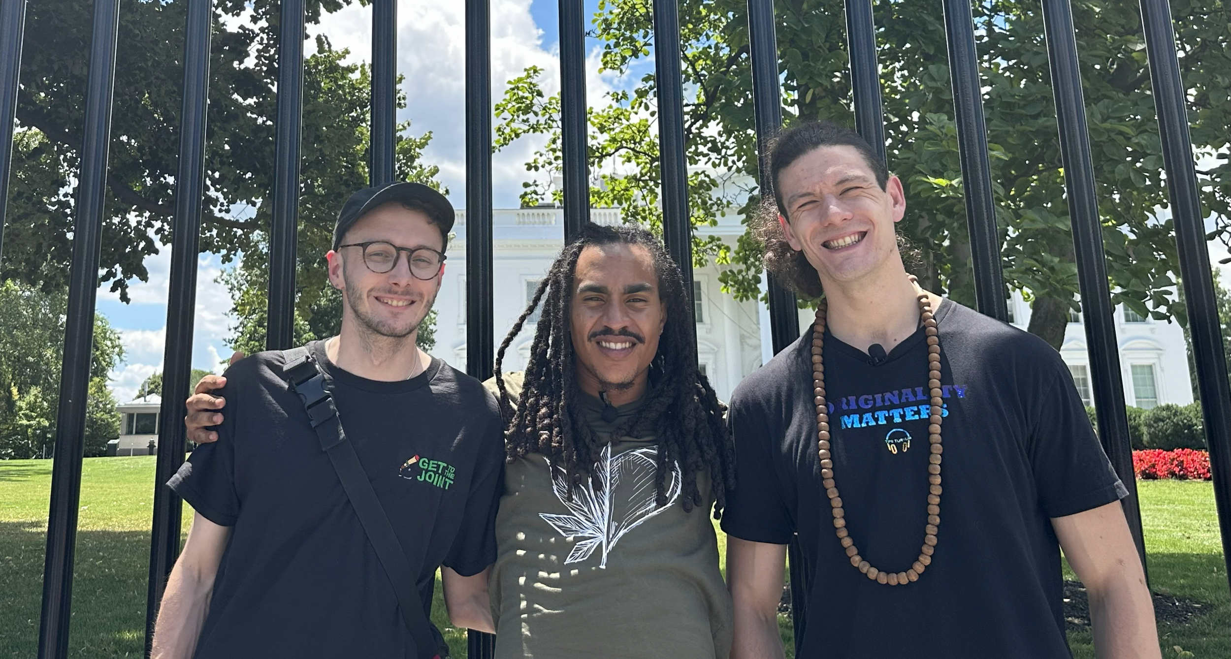 Three men standing outdoors in front of black fence, smiling, with the White House in the background on a sunny day.