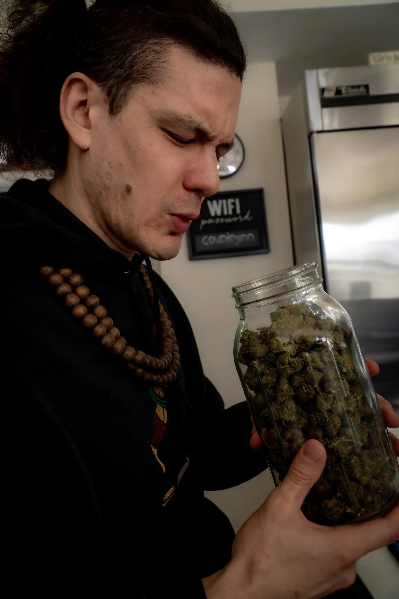 A person with dark hair and wearing a beaded necklace is holding a glass jar filled with dried cannabis buds. The person is looking at the jar with a focused expression. In the background, there's a sign indicating Wi-Fi password and a clock.