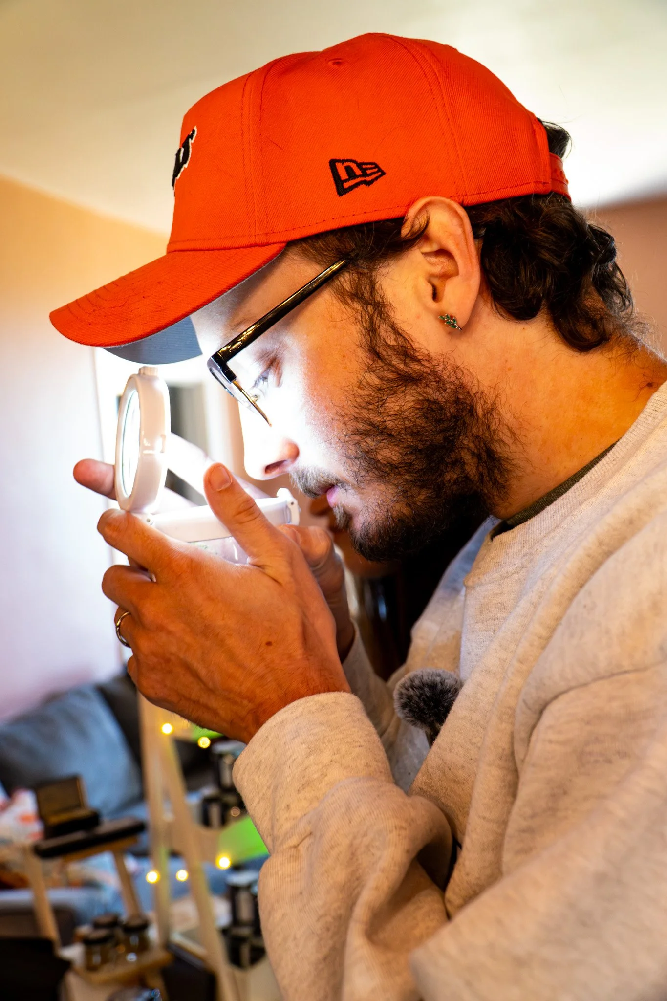 A man with glasses, a red cap, and a beard is holding a magnifying glass close to his face, examining something with focus. He is wearing earrings and a light-colored sweatshirt, with a background of a cozy room with shelves, chairs, and string lights.