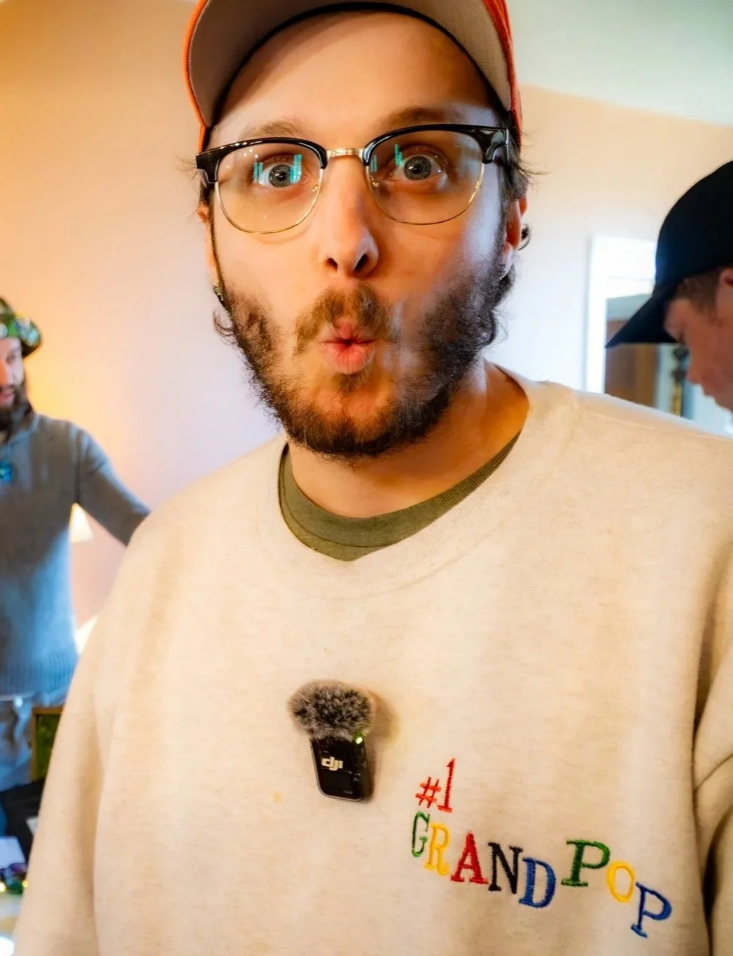 A man with glasses and a beard making a surprised face, wearing a beige sweatshirt with the word 'GRANDPOP' in colorful letters, a microphone clipped to his shirt, and a red cap. Two other men are visible in the background, one wearing a gray sweater and a green hat, and the other in a black cap.
