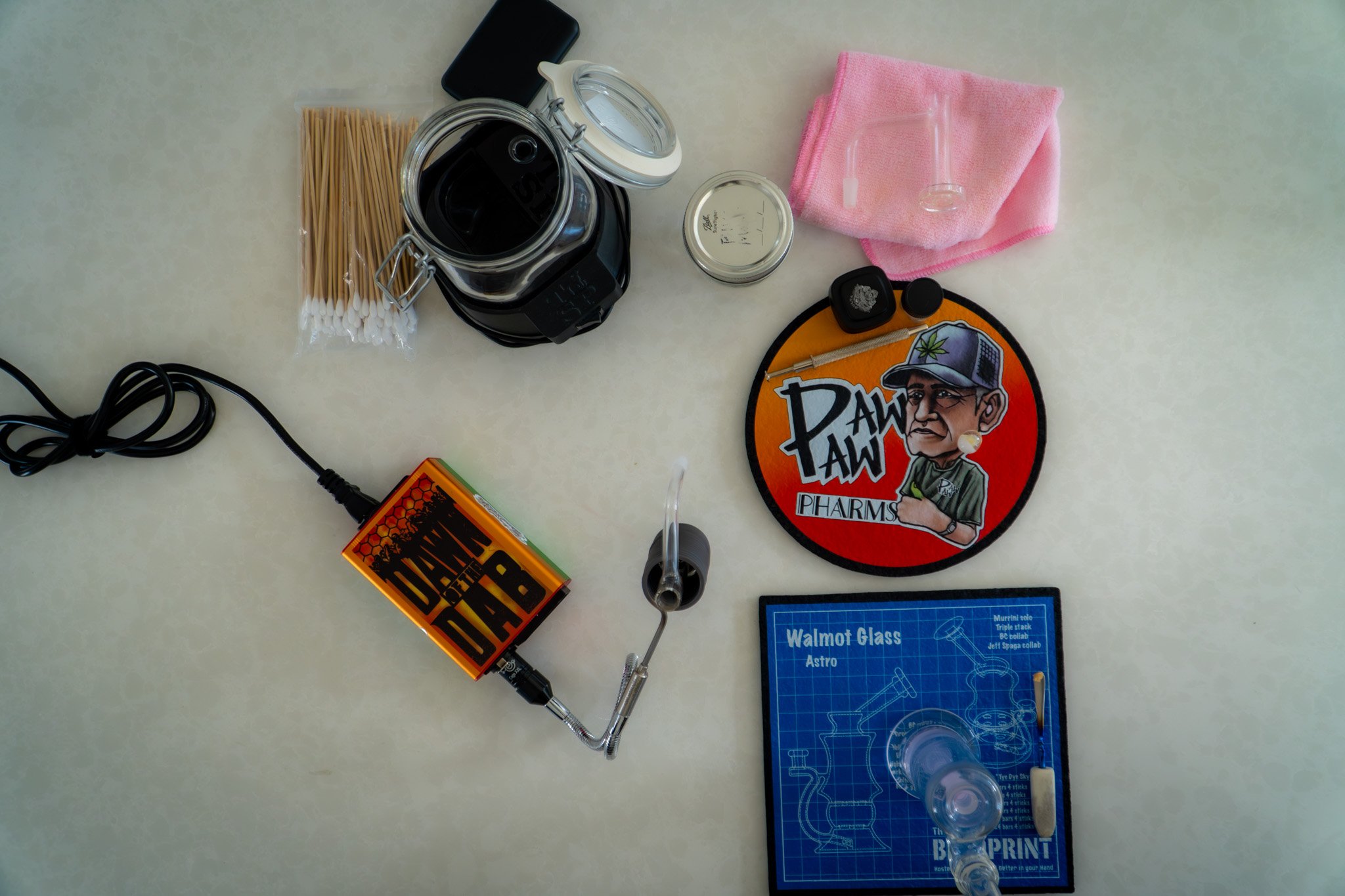 A collection of smoking accessories and paraphernalia on a table, including a glass bong, lighters, matches, a grinder, a pink cloth, a sticker, and a blue blueprint-style sheet labeled "Walmart Glass".