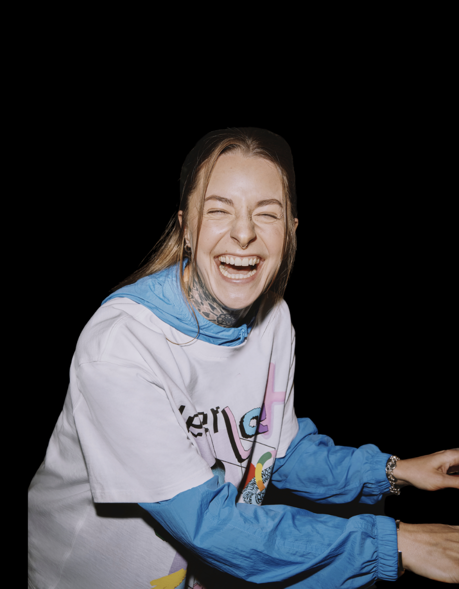 founder and president TJ with long, light brown hair, laughing with eyes closed, wearing a white graphic t-shirt with colorful designs and a blue windbreaker, against a black background