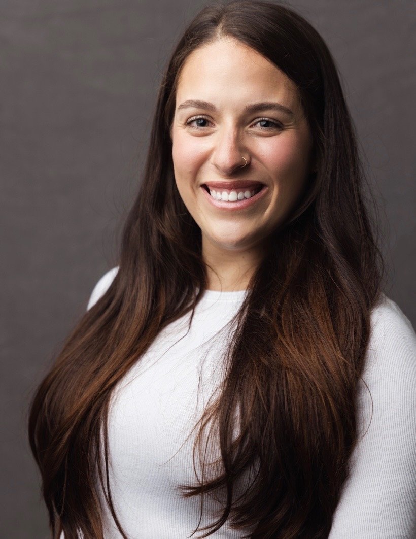 A young woman with long, wavy brown hair, smiling, wearing a white top, and standing against a gray background.