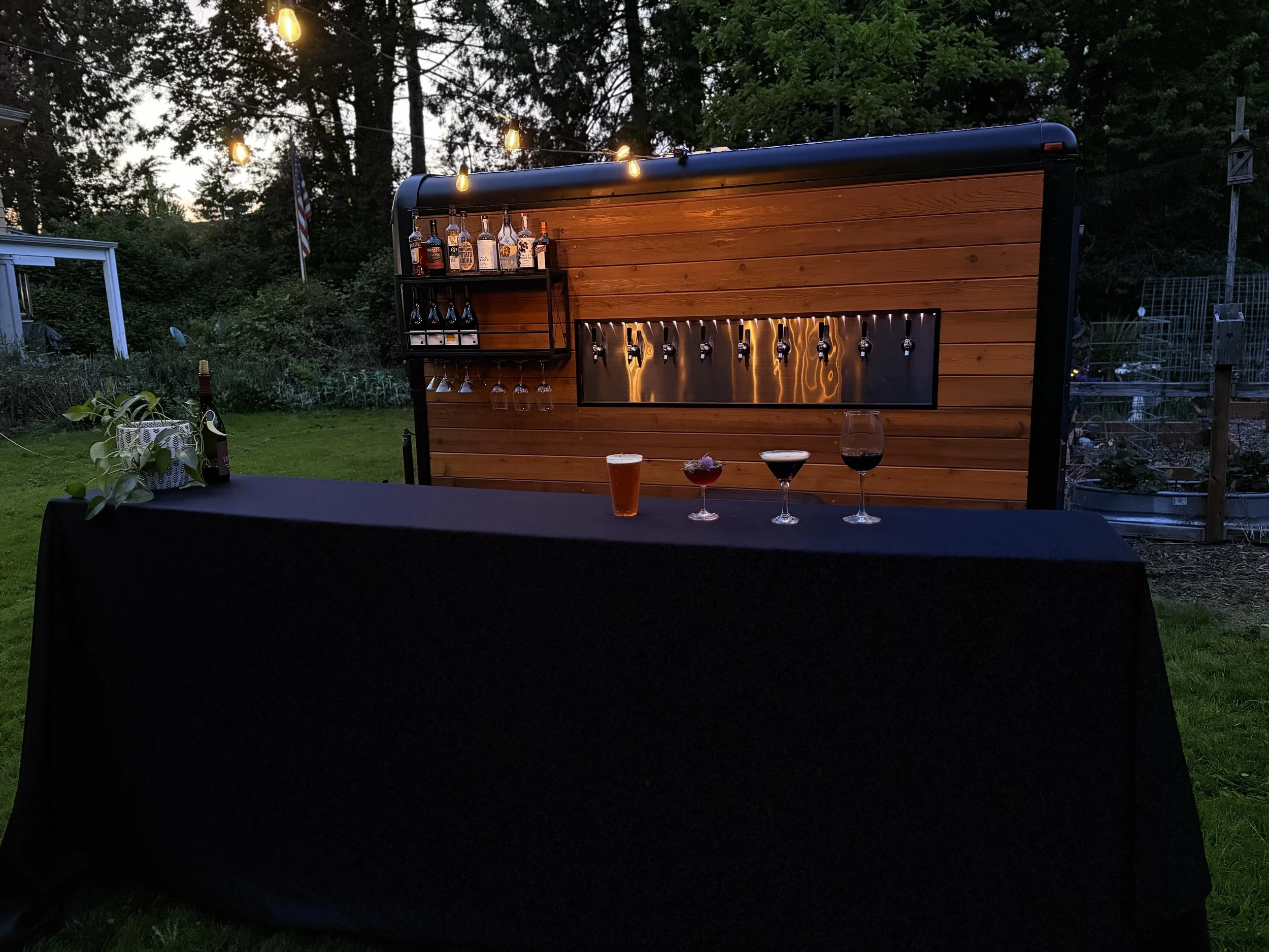 An outdoor bar setup at dusk with bottle display, hanging wine glasses, and four different cocktails on a black table, with string lights and trees in the background.