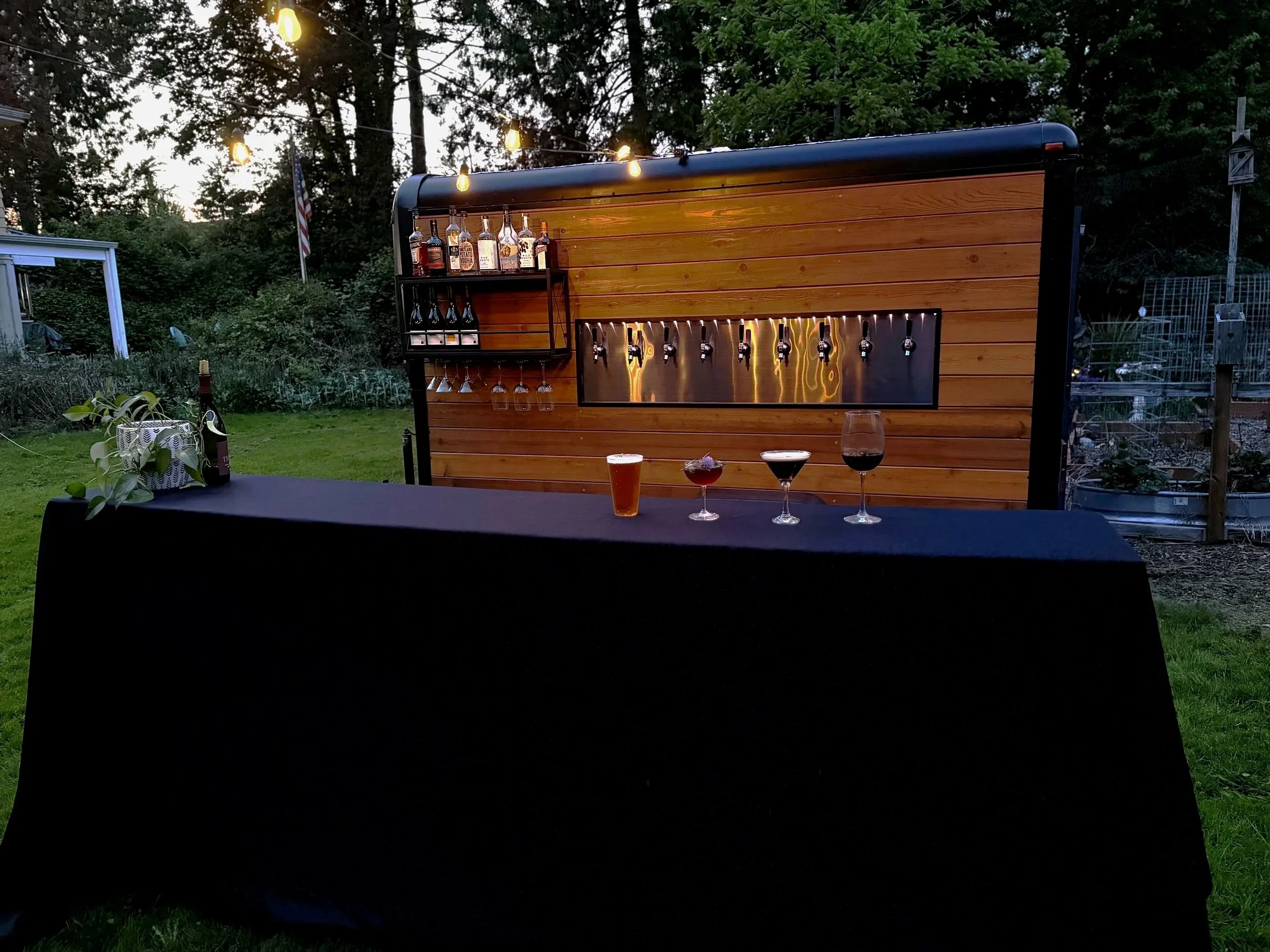 Outdoor backyard scene with a wood-paneled mobile bar setup decorated with string lights. Three glasses with beverages and a wine glass are on a black table in the foreground. There are bottles of liquor on the bar, and the background features green 