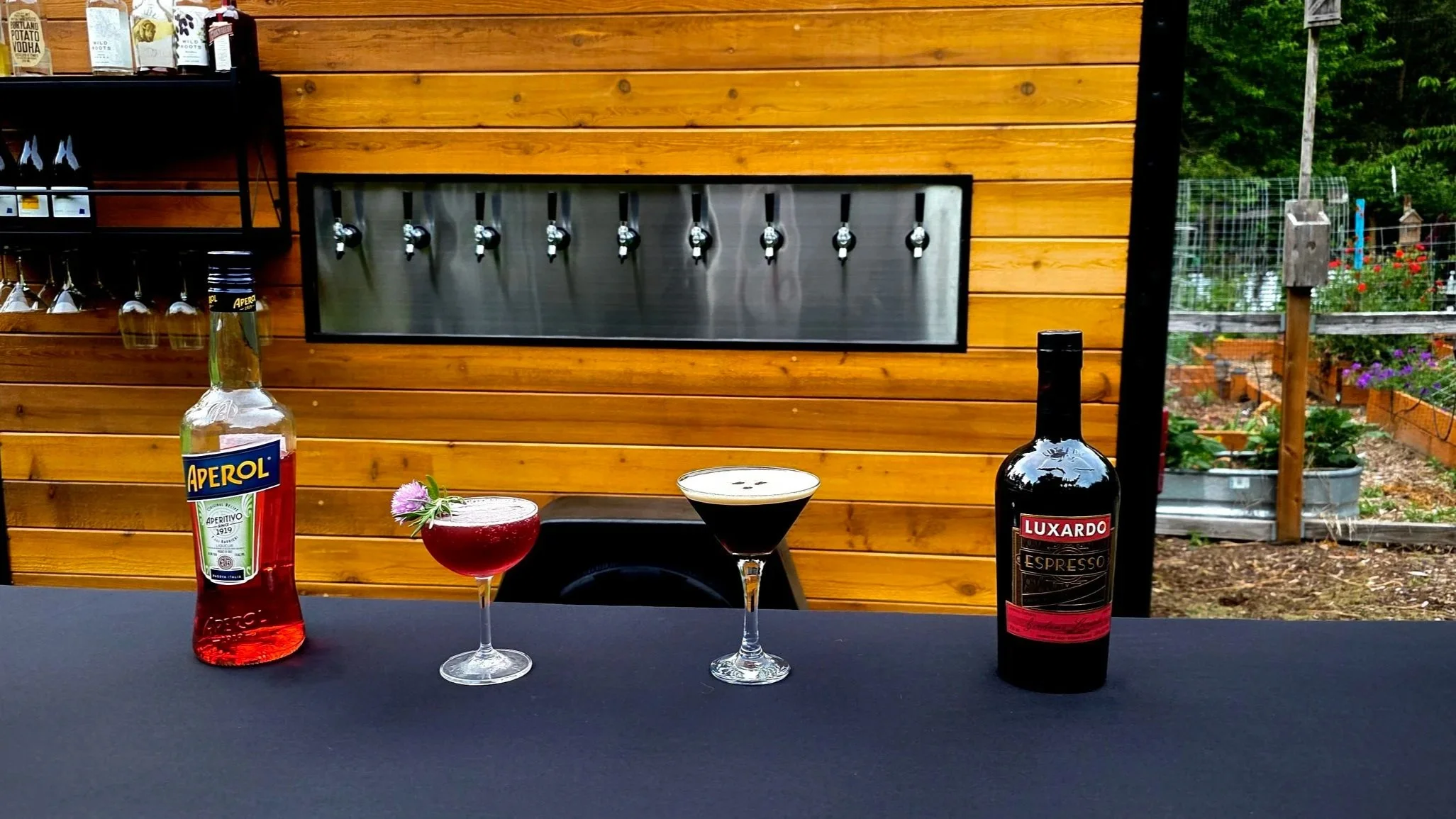 A wooden outdoor bar area with various bottles, including Aperol and Luxardo Espresso, on the counter. There are three cocktails on the table: one red drink with a flower garnish, a dark cocktail in a martini glass, and a frothy drink in a glass with