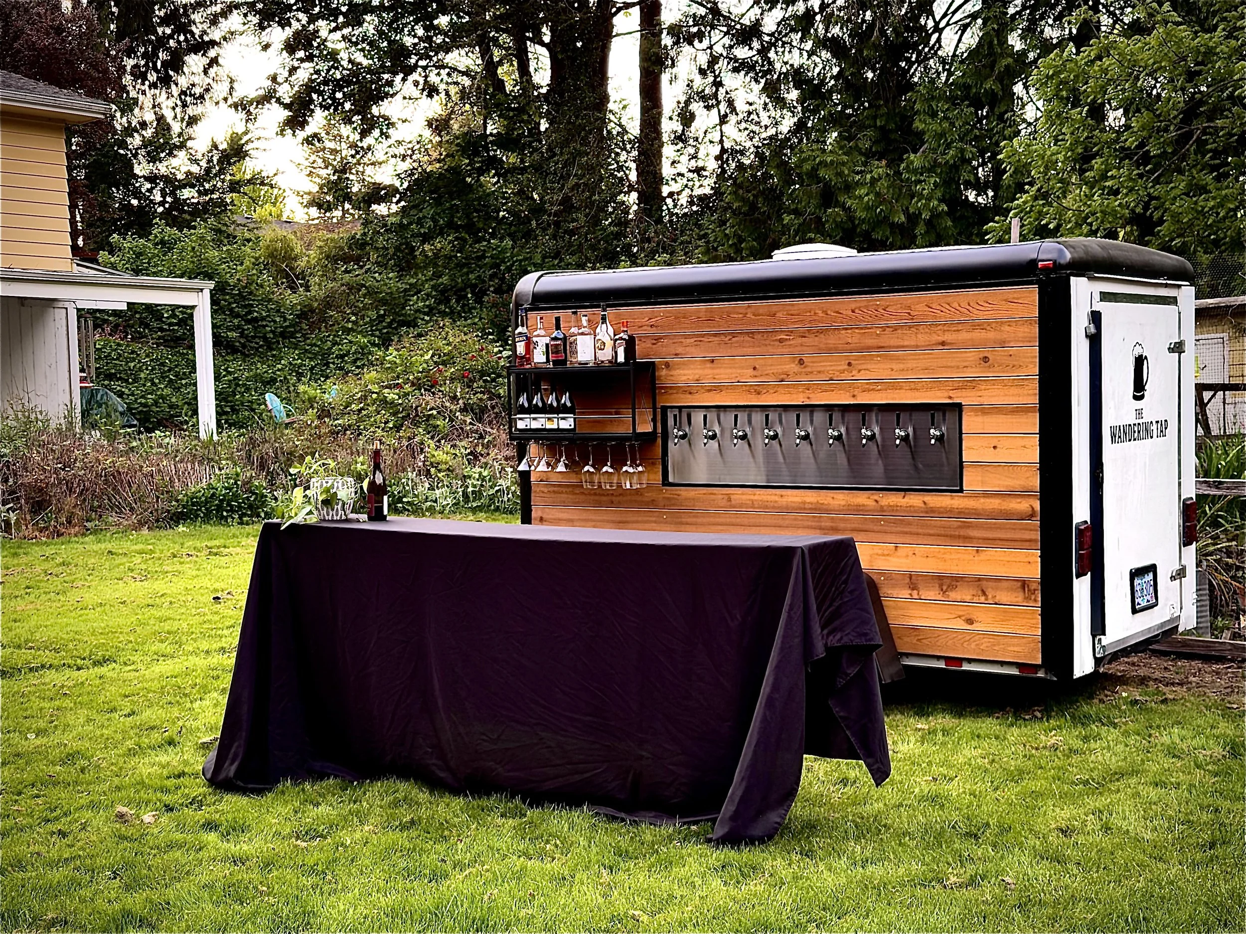A mobile bar setup with several bottles of alcohol on top of a wooden structure behind a black tablecloth covered table outdoors, with trees and a house in the background.