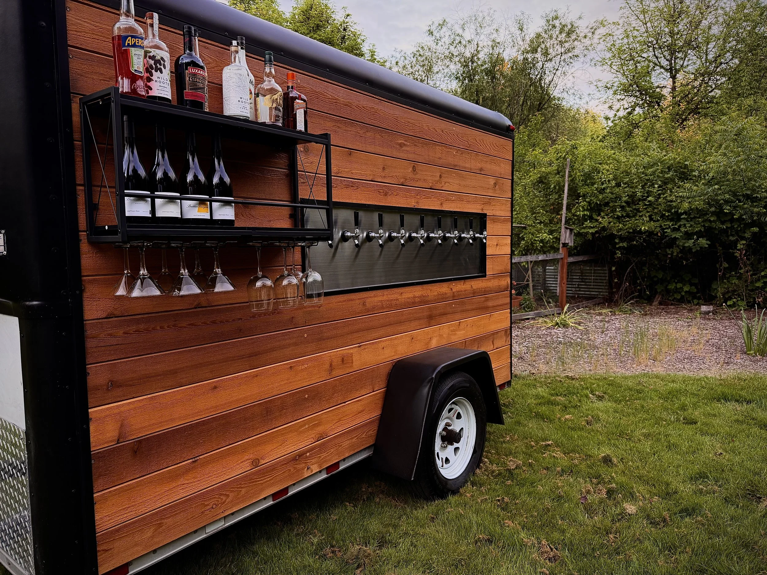 A mobile bar trailer with a wooden exterior featuring an outdoor alcohol and wine station. The station has several bottles of alcohol, wine glasses hanging upside down, and taps for dispensing drinks. The trailer is parked on a grassy area with trees