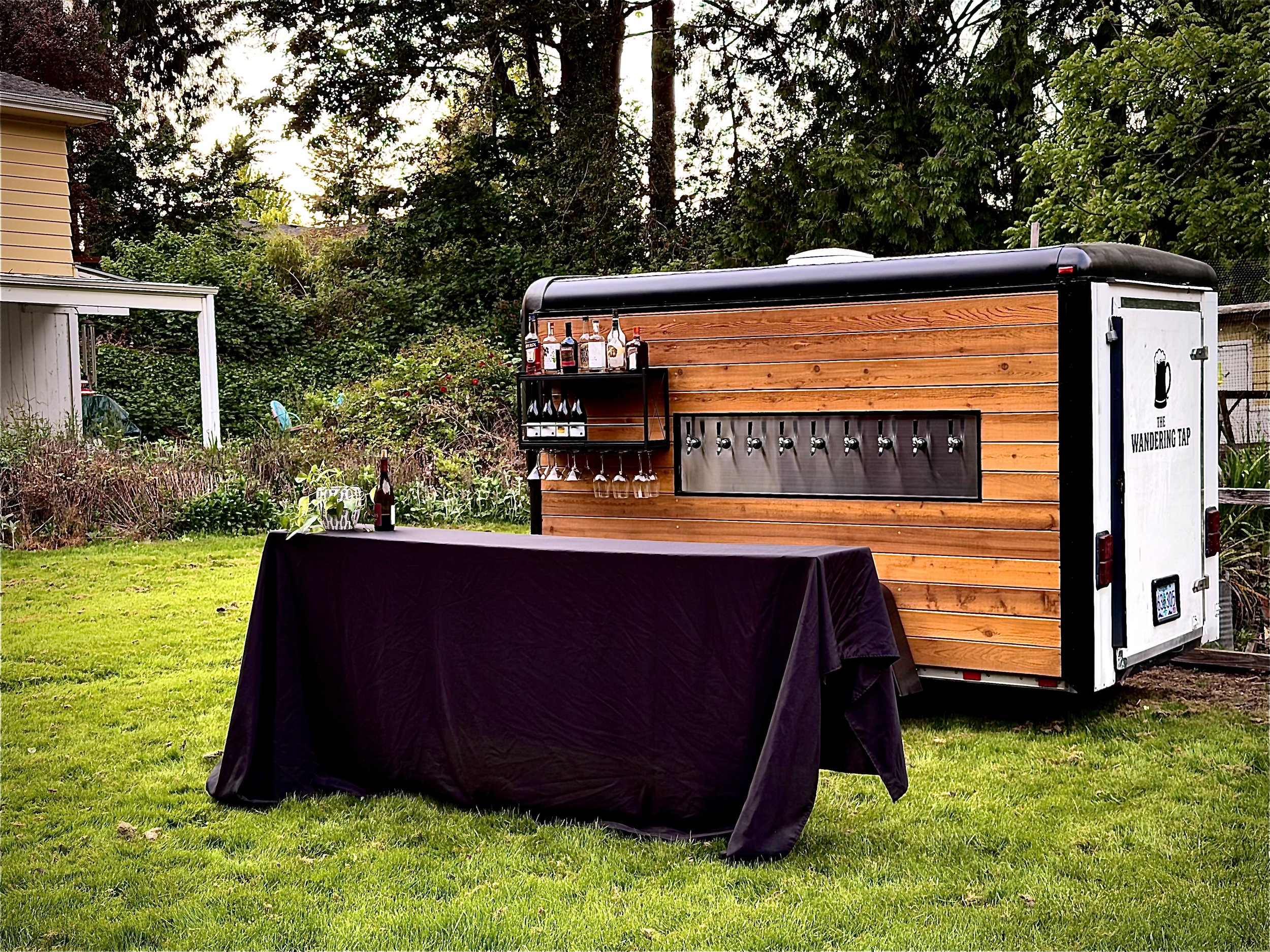 A mobile bar setup on a trailer with a wooden exterior, featuring various bottles of alcohol on shelves, set in a backyard with lush green trees and grass.