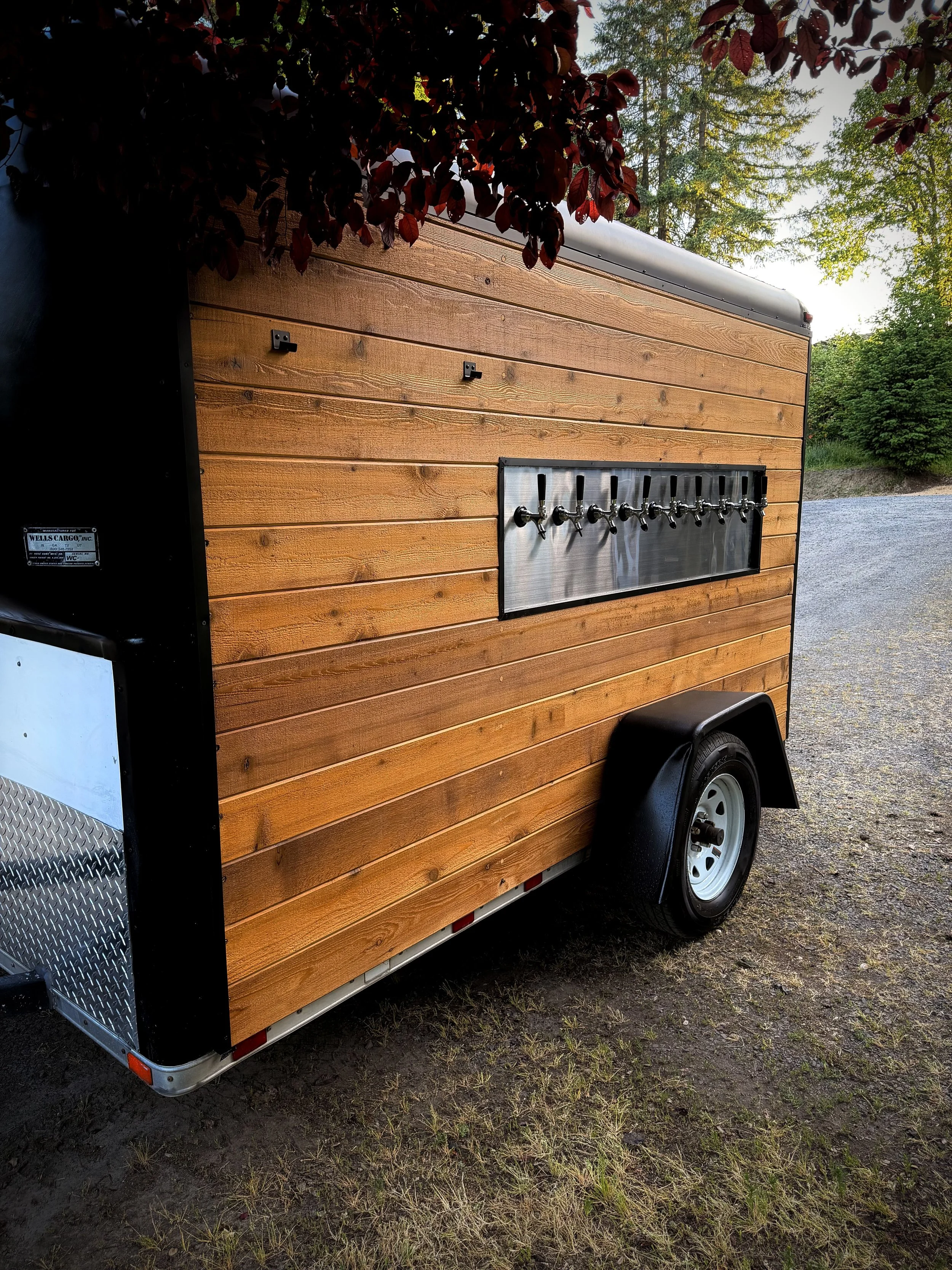 A food and beverage trailer with a wooden exterior, equipped with multiple beverage taps, parked on a gravel and grass surface surrounded by trees and greenery.