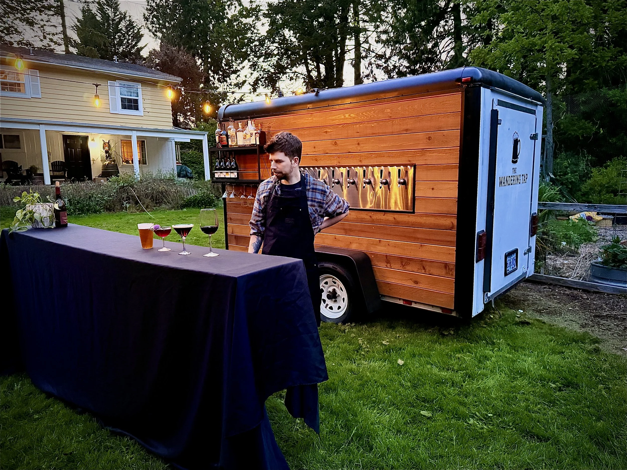 A man standing next to a small mobile bar called 'The Wandering Tap' set up outdoors in a backyard, with a table in front holding glasses of drinks, and trees and a house in the background.