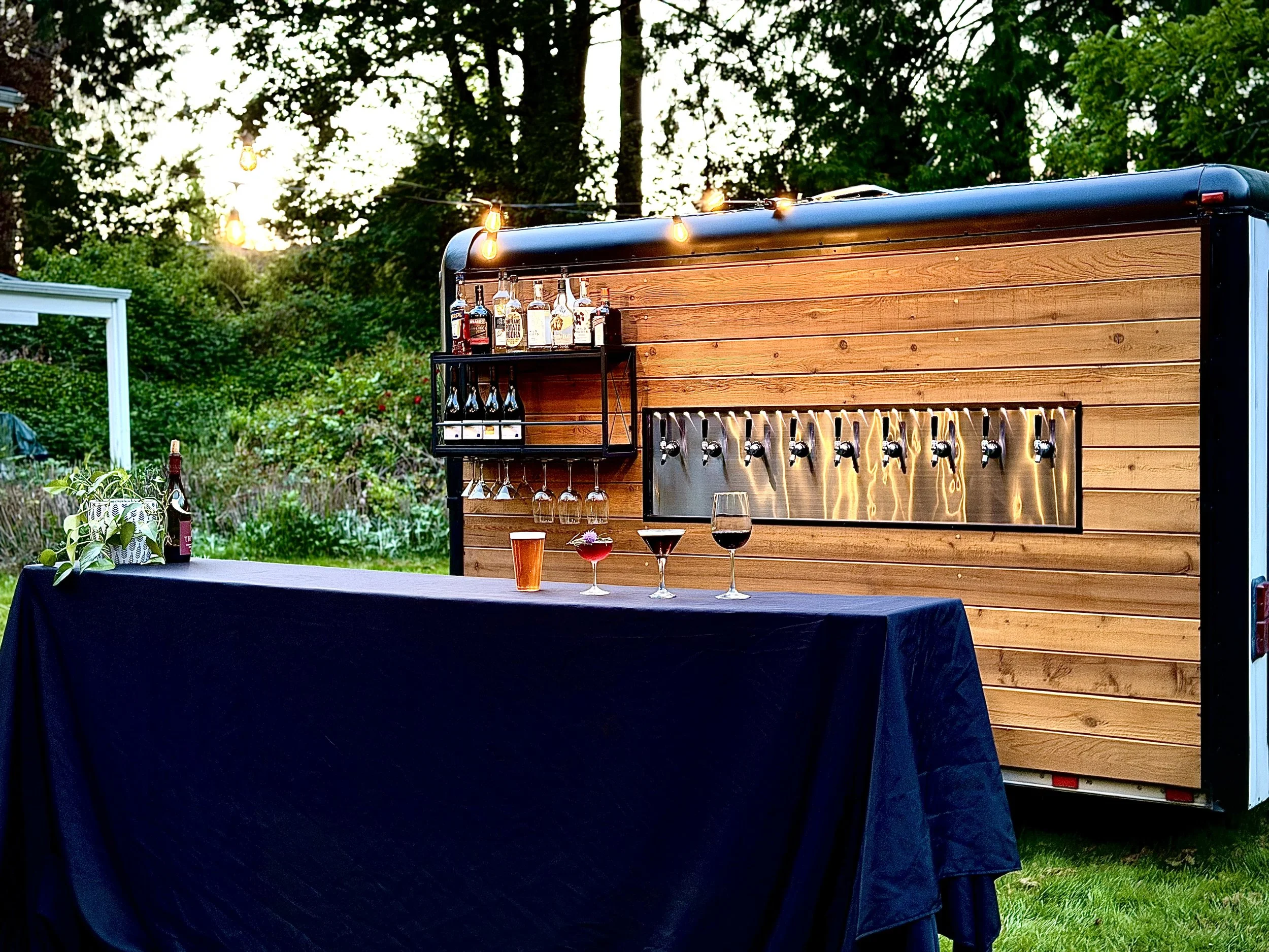 Outdoor bar setup with liquor bottles and glasses on a table, with string lights hanging overhead and a wooden backdrop.
