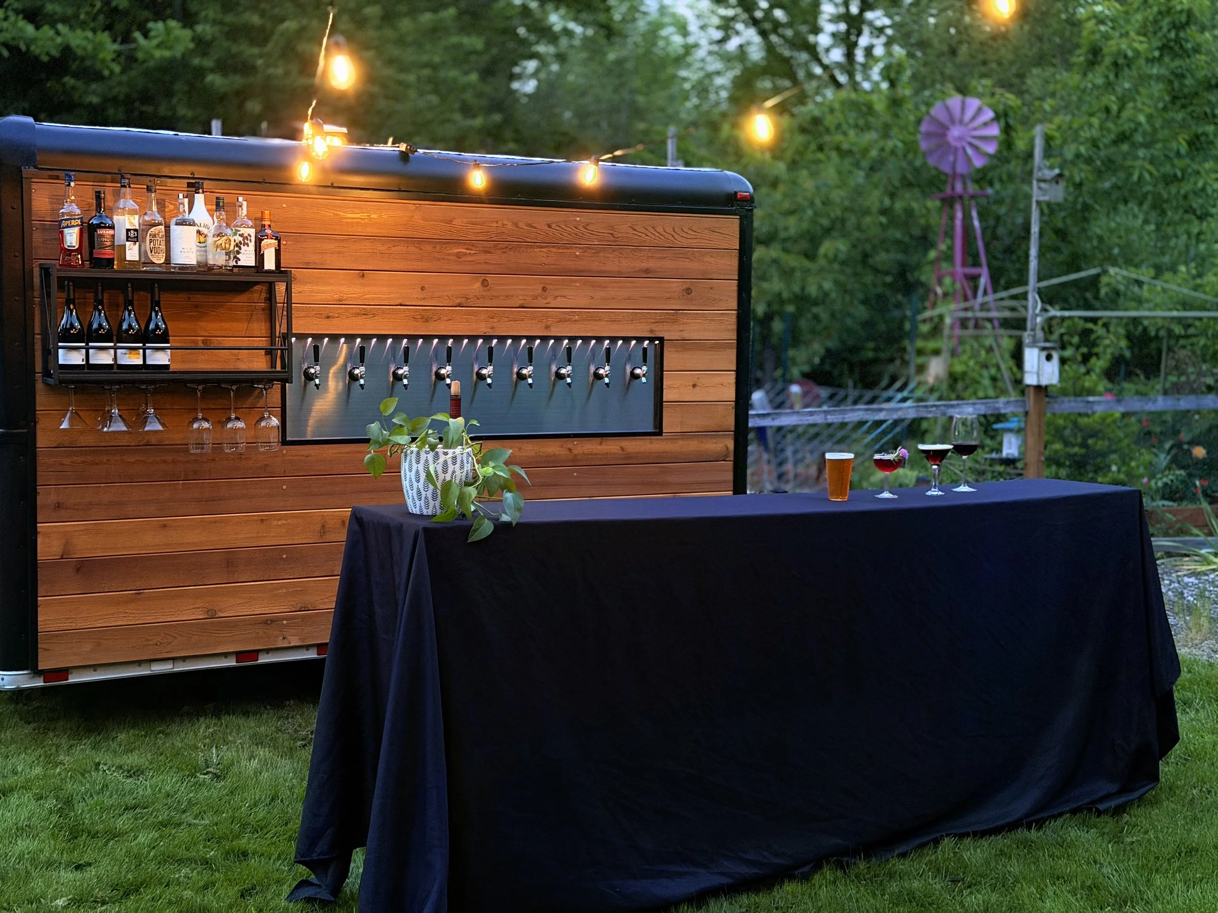 Outdoor bar setup with liquor bottles on a wooden trailer and glasses hanging underneath, decorated with string lights and a table with drinks and a potted plant, surrounded by greenery and a windmill in the background.