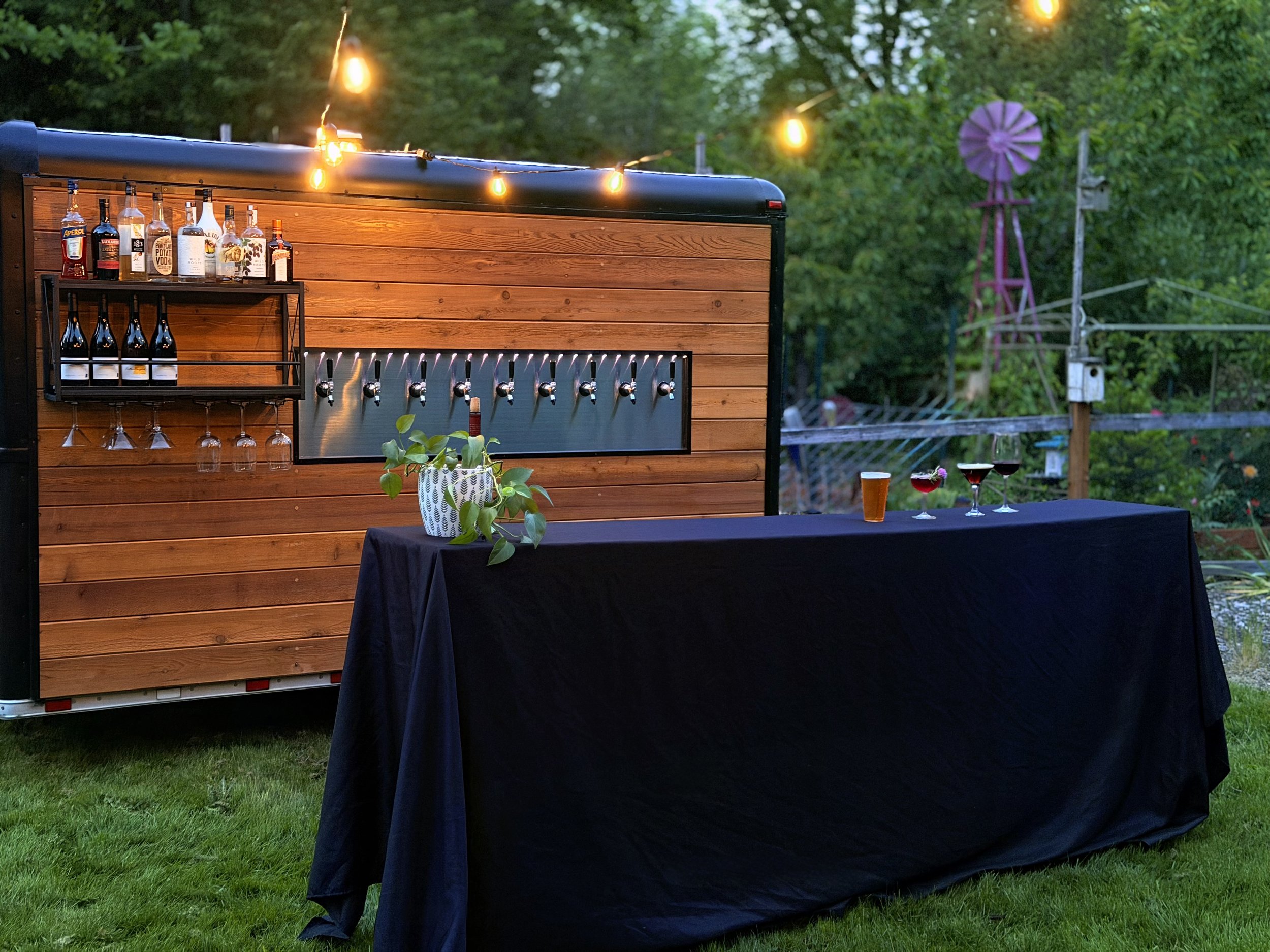 Outdoor scene showing a food or drink trailer with string lights, bottles, and glasses, set up on a grassy area. There is a table with a black tablecloth, red and clear drinks, and a potted plant. In the background, trees and a windmill are visible.