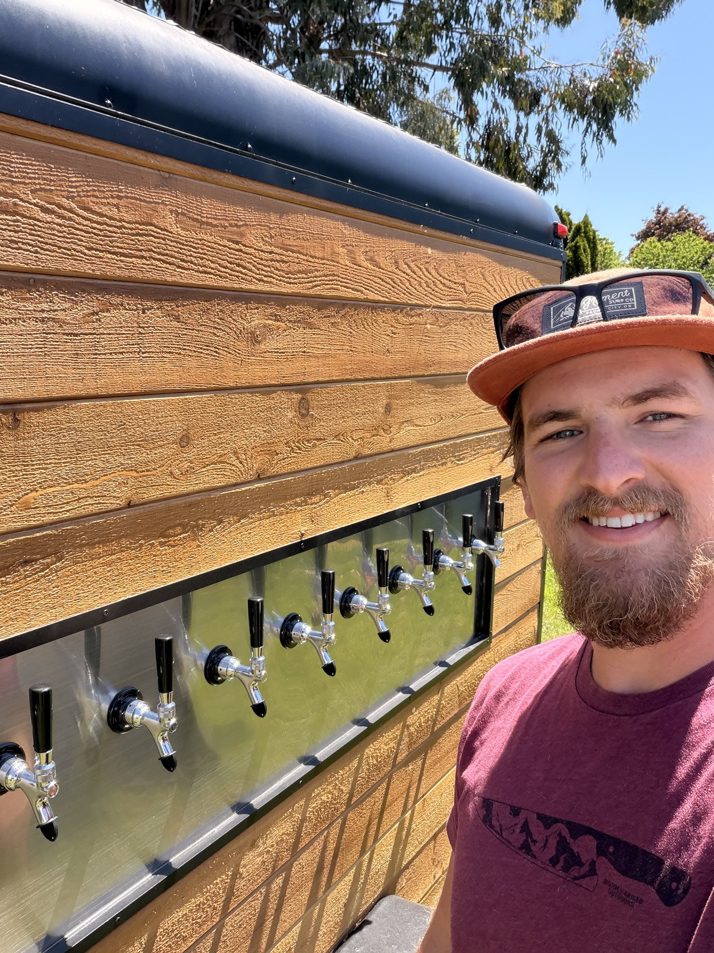A man with a beard and a maroon shirt taking a selfie outdoors. Behind him, there is a wooden-paneled wall with a stainless steel beverage dispenser with multiple taps. The man is wearing sunglasses on his hat.