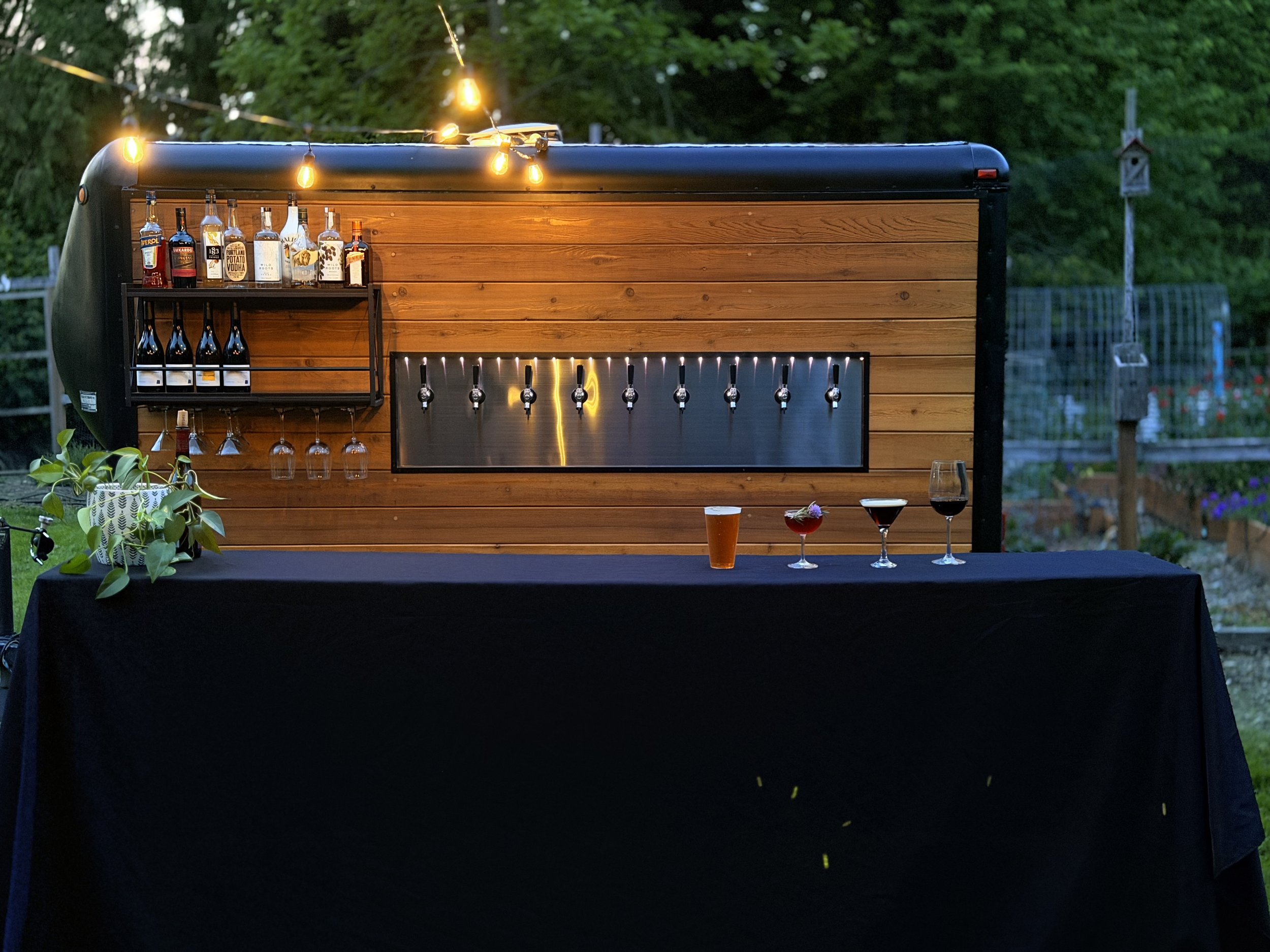 Outdoor bar setup with liquor bottles, glassware, and a row of beer taps, decorated with string lights in a backyard setting.