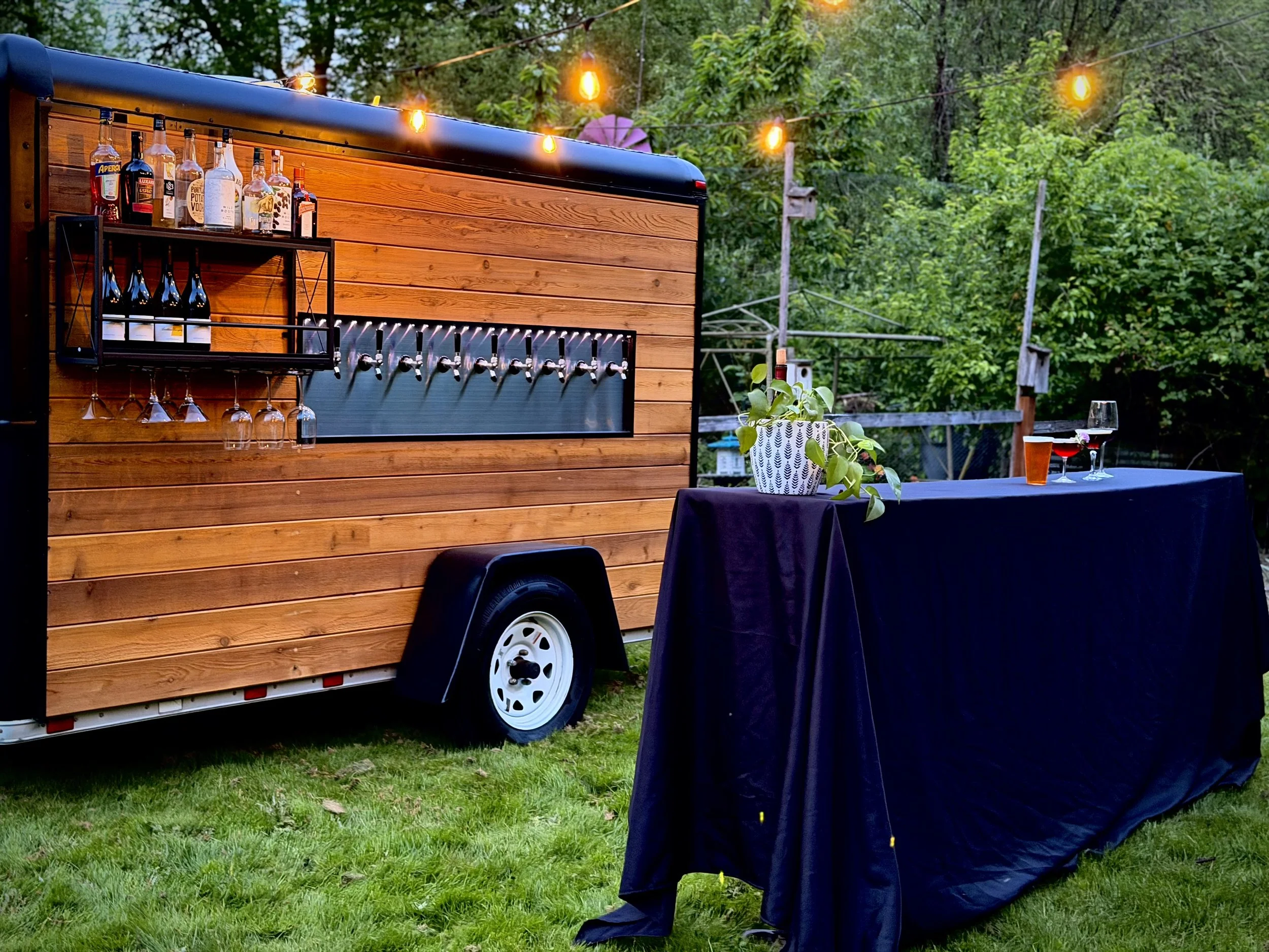 A mobile bar on wheels serving drinks at an outdoor event, with a table covered in a dark cloth, potted plant, and glasses of beer and red wine in the foreground. String lights hang overhead, and a lush green background is present.