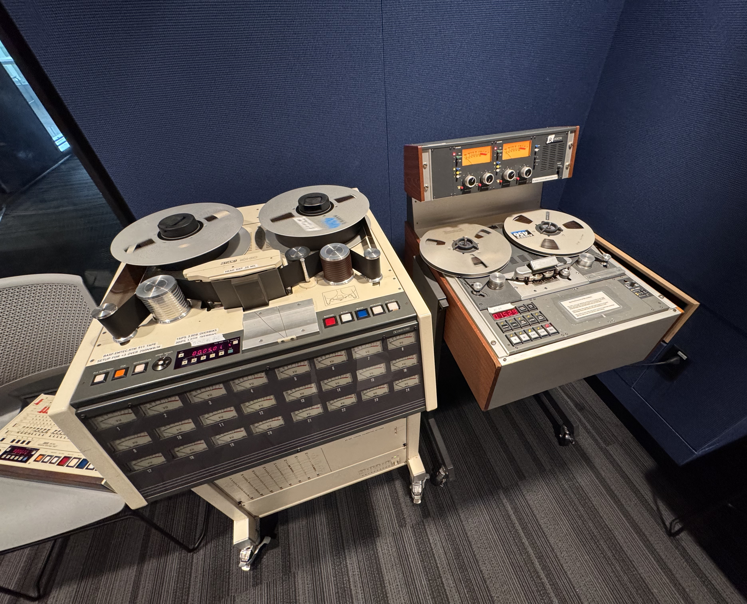 A vintage reel-to-reel tape recorder on a stand next to an audio processor with knobs and meters, in a room with blue acoustic wall panels and carpeted floor.