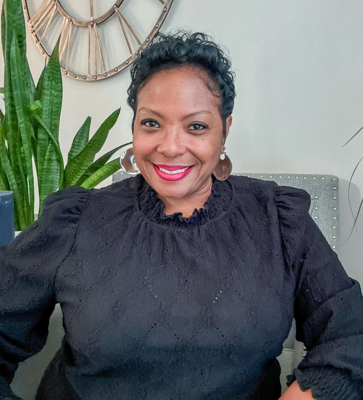 A woman with short curly black hair, wearing a black textured blouse and silver earrings, smiling while seated in an indoor space with a large green plant and a decorative wall clock in the background.