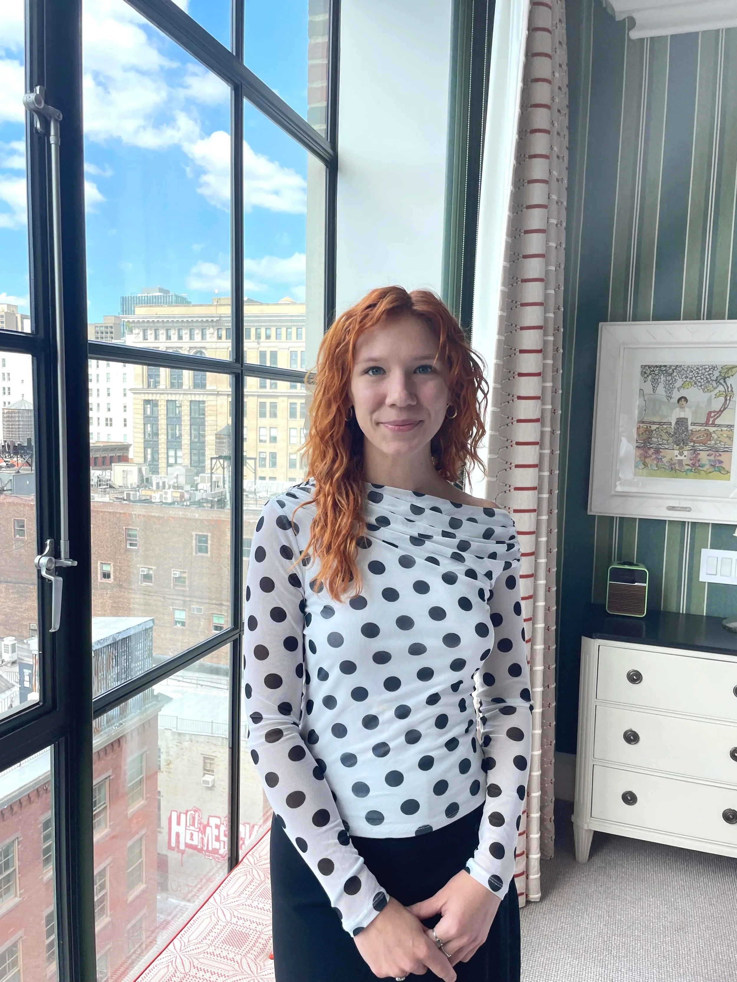 A woman with wavy orange hair, wearing a white long sleeve top with black polka dots, standing indoors near a large window with city buildings outside.