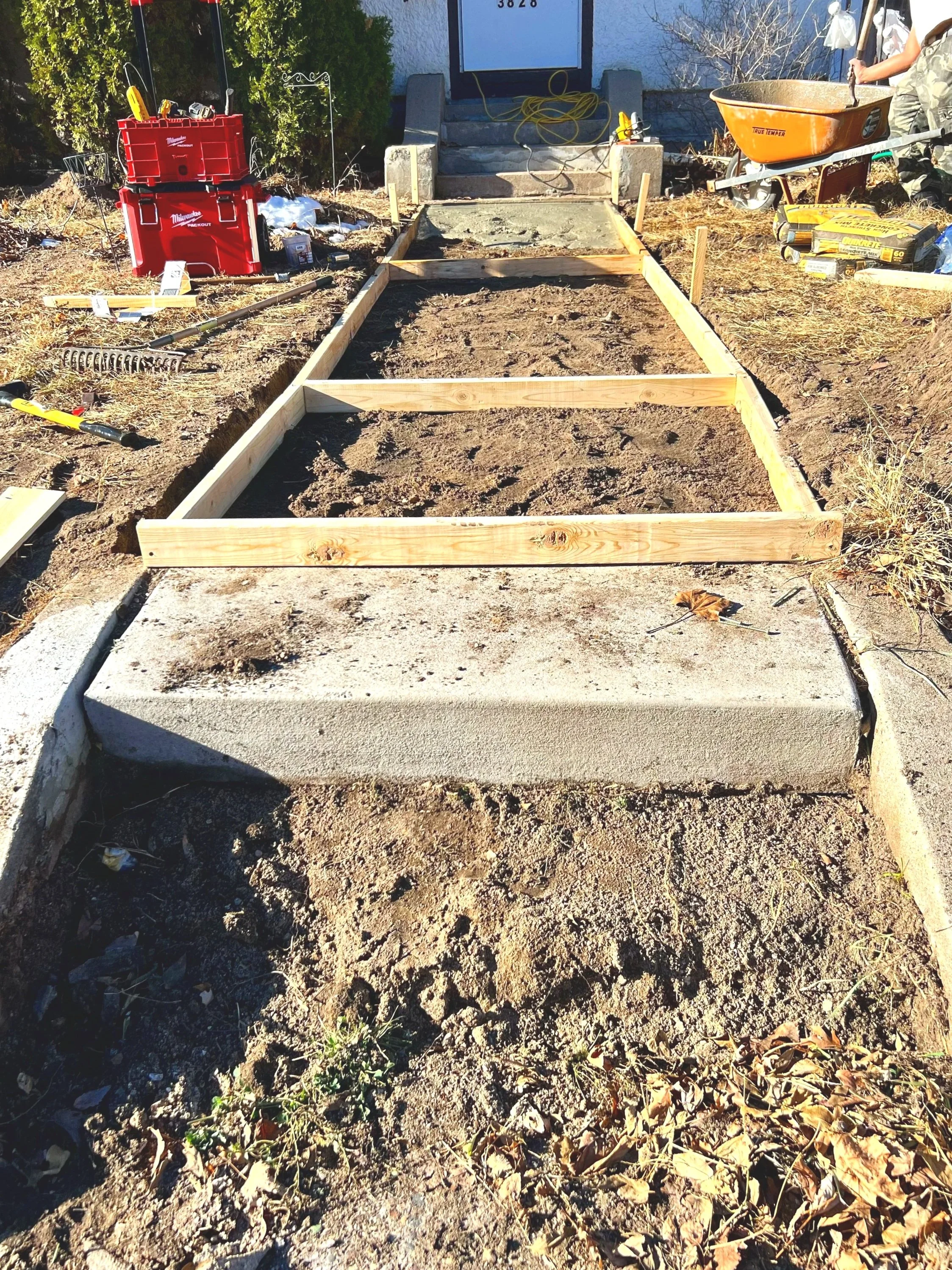 A construction site showing the beginning of a concrete sidewalk or patio with wooden framing. Construction tools and supplies are visible, including a wheelbarrow, red storage container, rake, and packaging for concrete or cement mix. A person holds